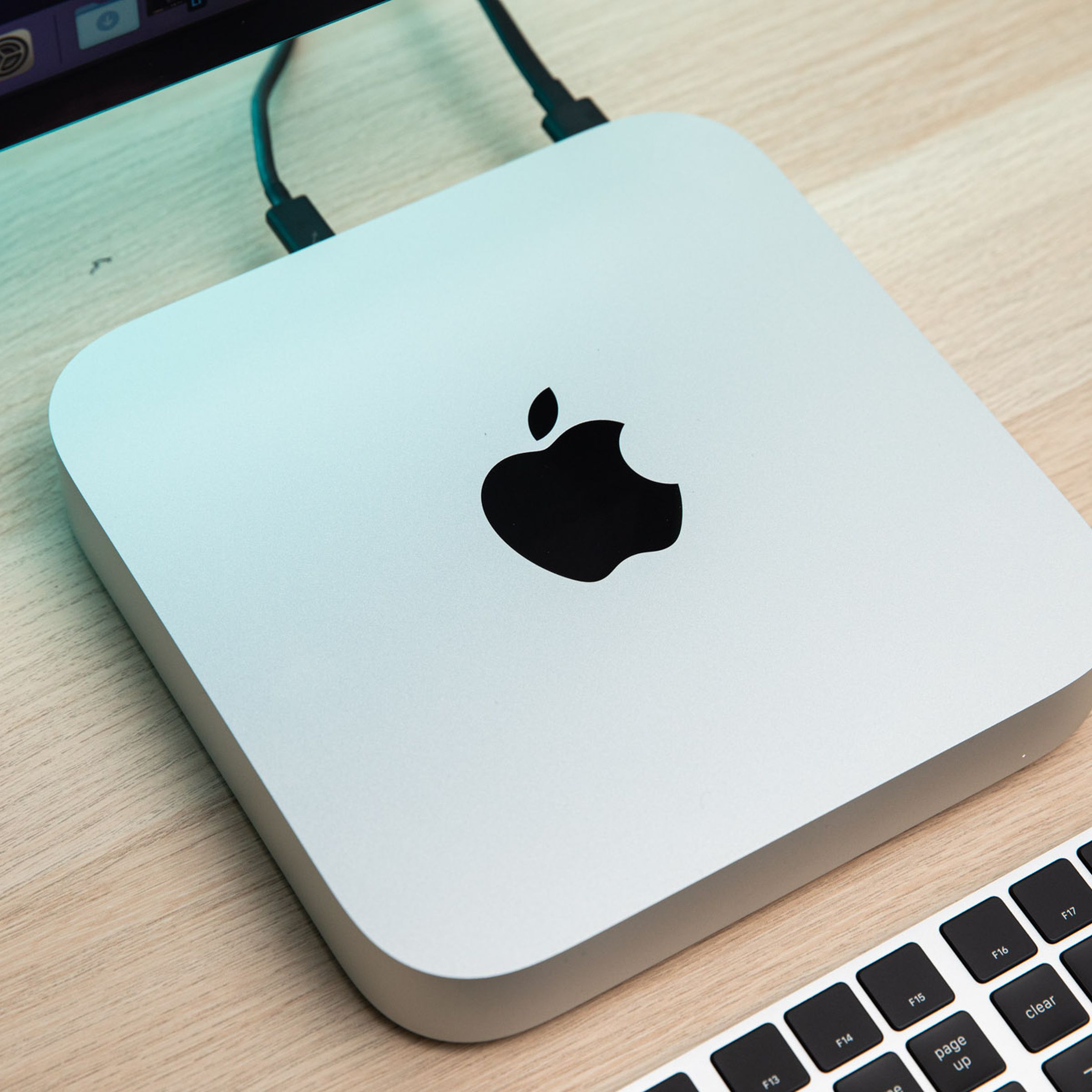 The Mac Mini sitting on a wood table underneath a monitor and beside a keyboard.