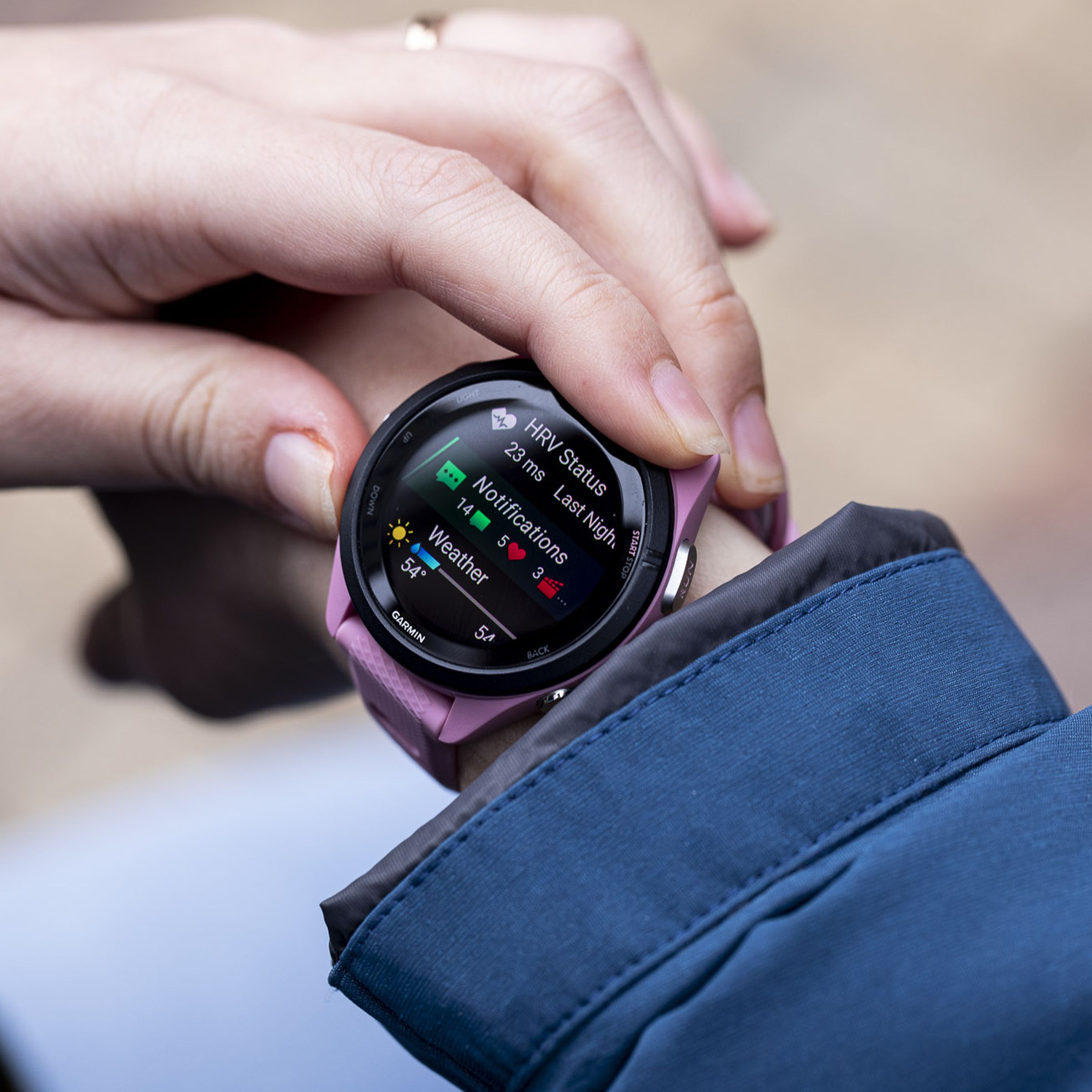 A close-up image of a pink Garmin Forerunner 265S on a woman’s wrist, just outside a blue raincoat.