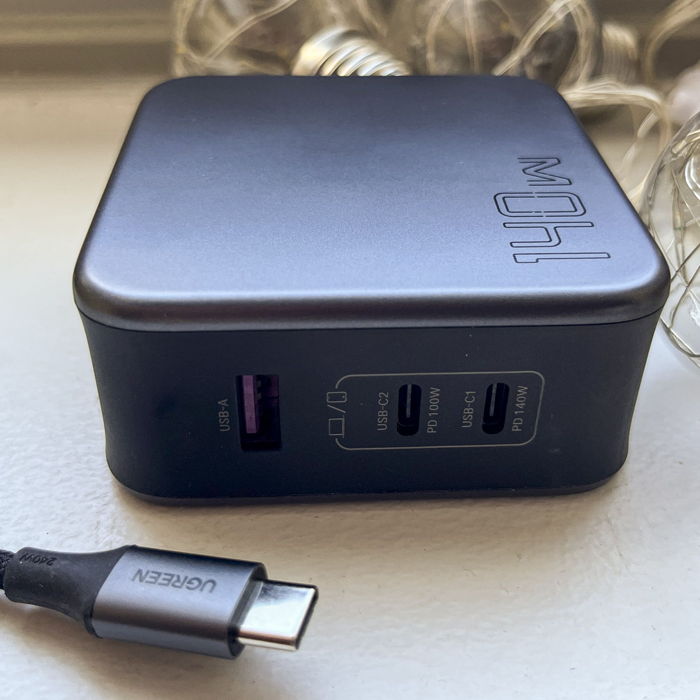 Ugreen’s Nexode charger is a gray brick on its side on a white window sill. 140W is printed on the top, string light retro bulbs sit behind it, and a braided black USB-C cable with Ugreen is printed on the tip. The charger has two USB-C ports and one USB-A port.
