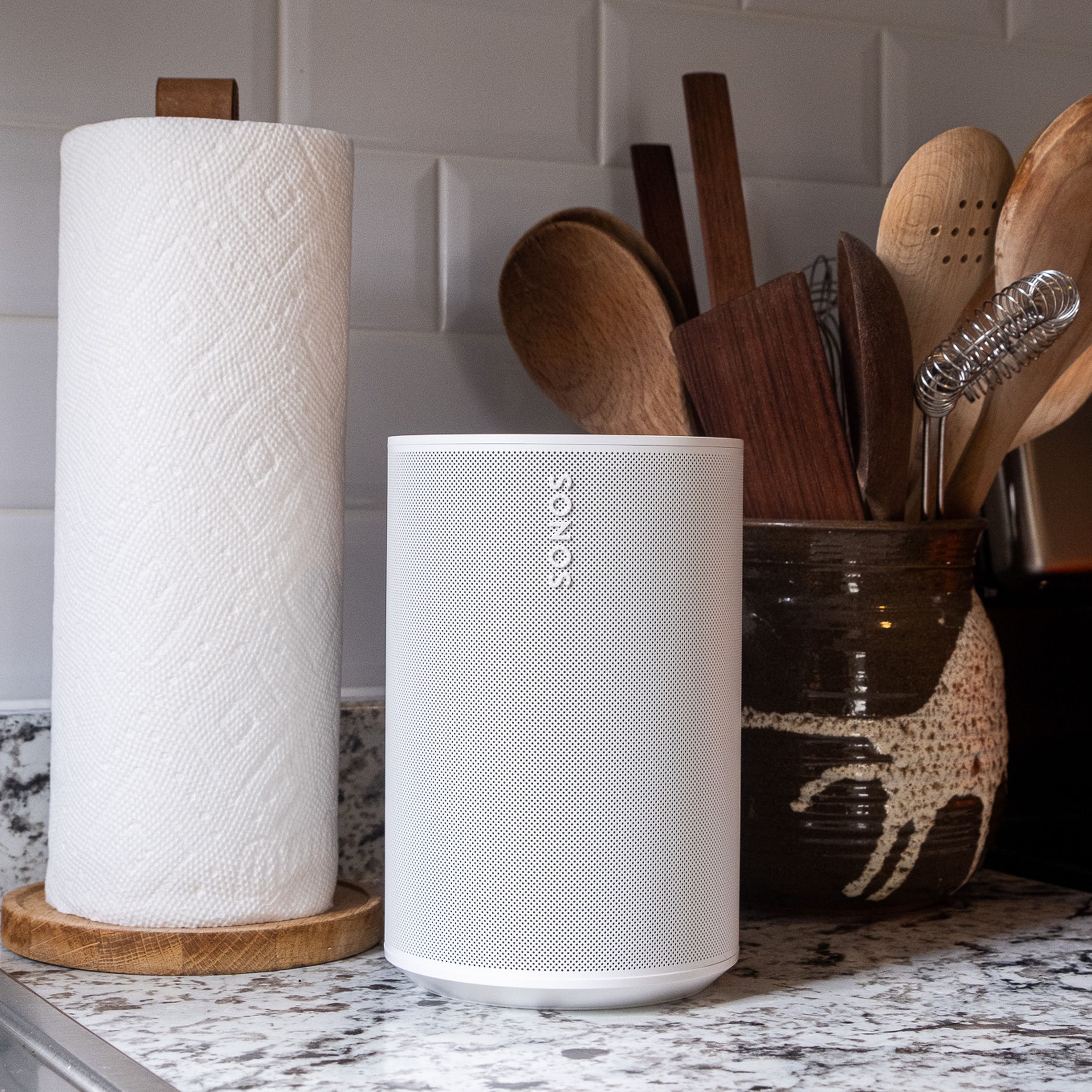 A white Sonos Era 100 speaker sitting on a marble kitchen countertop beside a paper towel roll and cooking utensils.