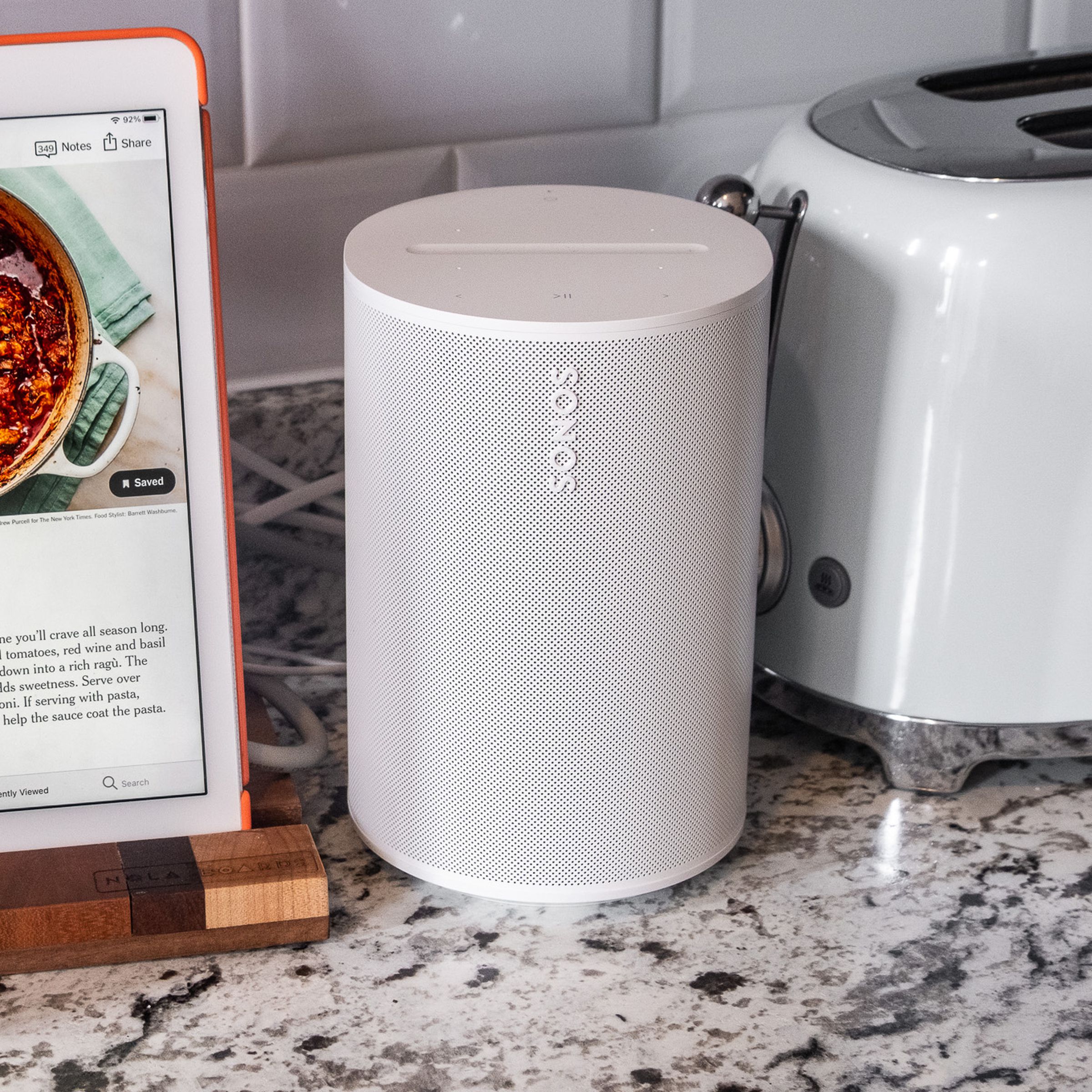 A photo of the Sonos Era 100 speaker in a kitchen setting beside an iPad and toaster.