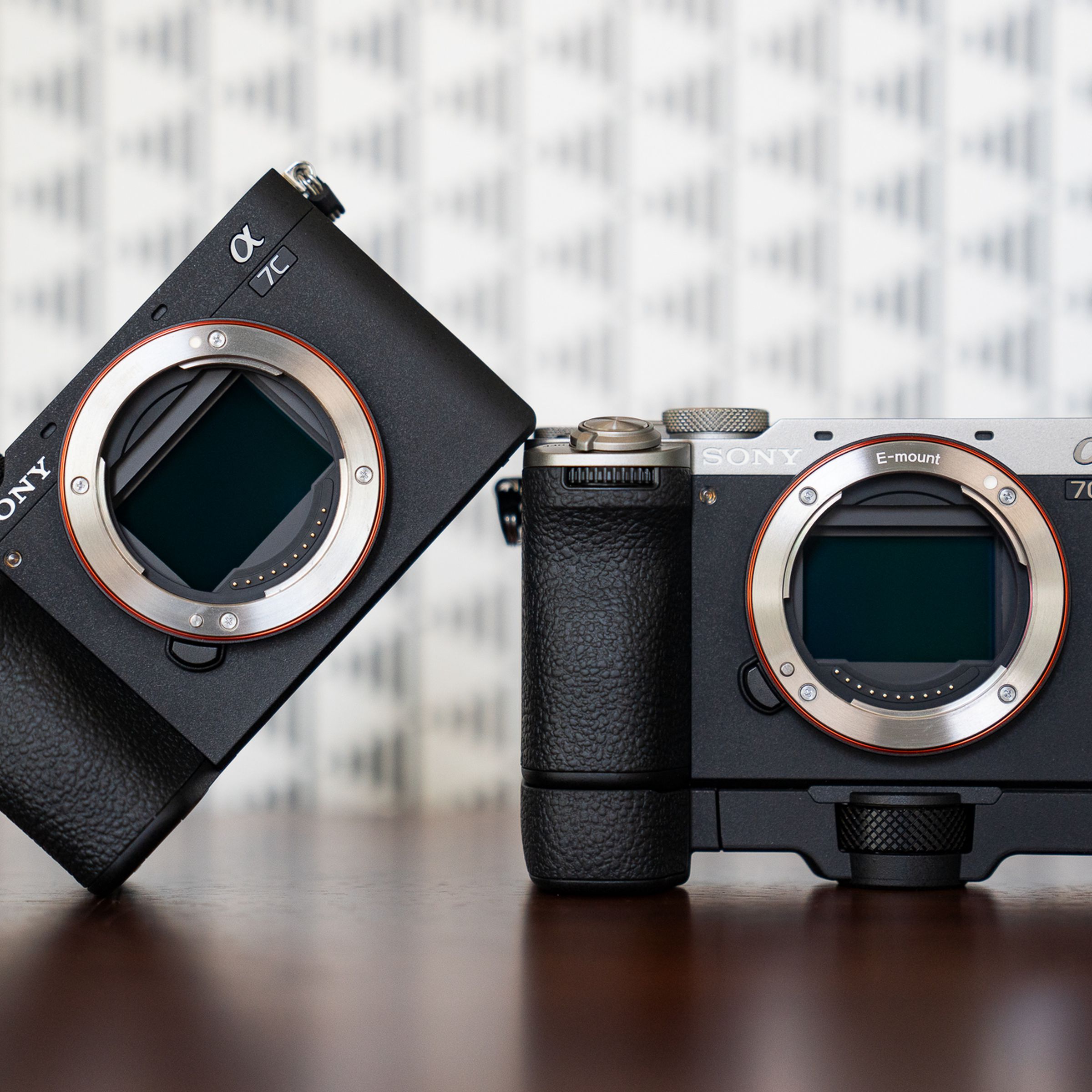 The Sony A7C II and A7C R cameras, sitting on a wood surface without lenses attached. The A7C II in black is angled and resting one side on the silver / black A7C R, which is mounted to its own handgrip.