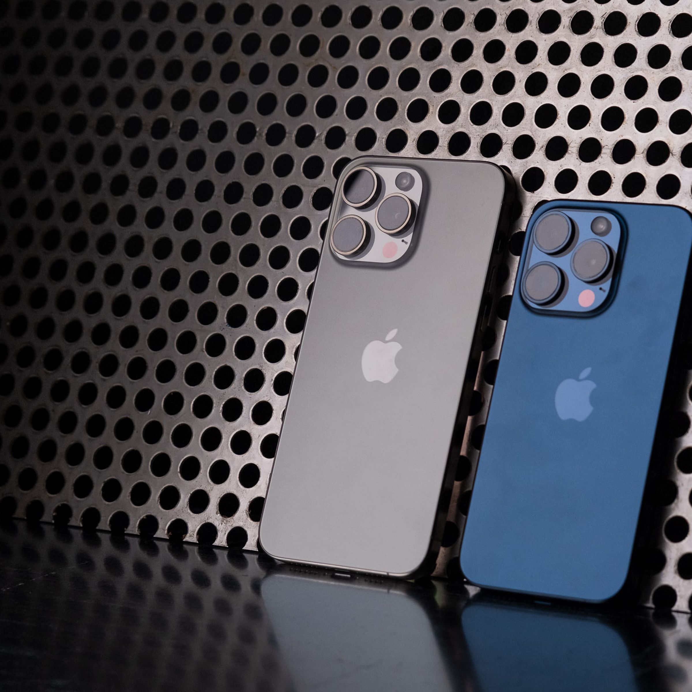 Blue iPhone 15 Pro and natural titanium iPhone 15 Pro Max leaning against a perforated metal background.