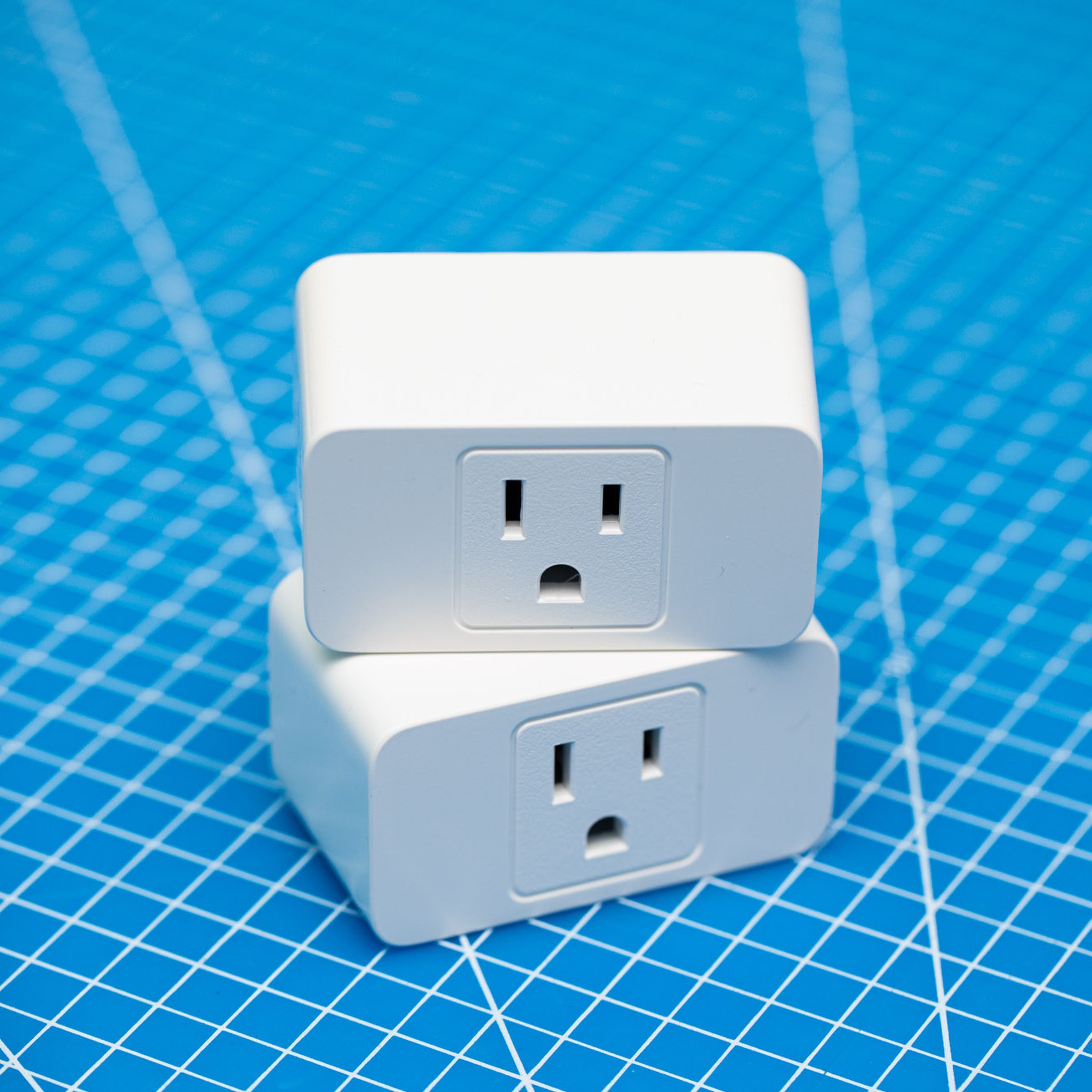 Two white Meross smart wi-fi plugs stacked on top of each other on a blue background. Each has a three-prong outlet on the front face.