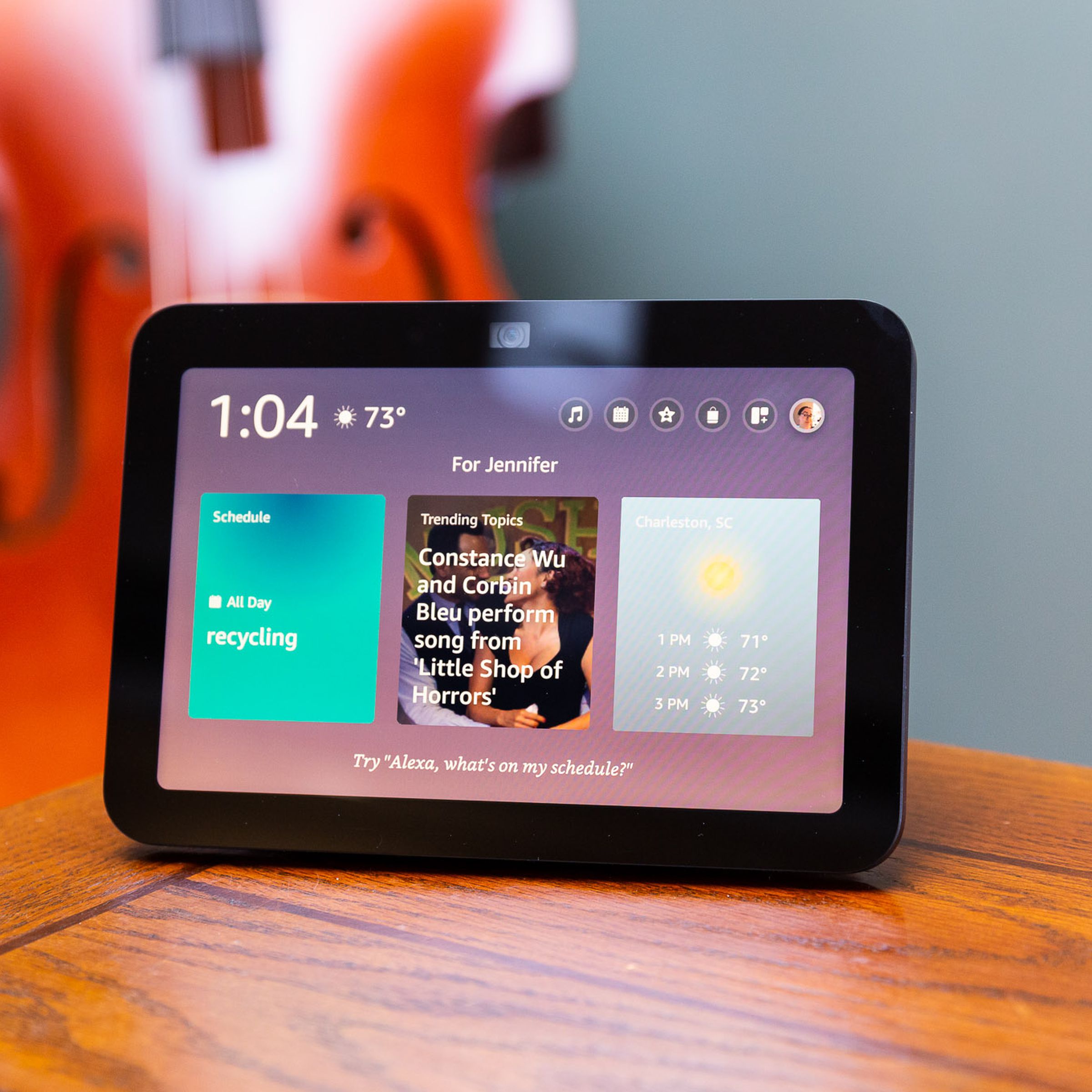 An Echo Show smart display on a wooden table with a wooden bass in the background.