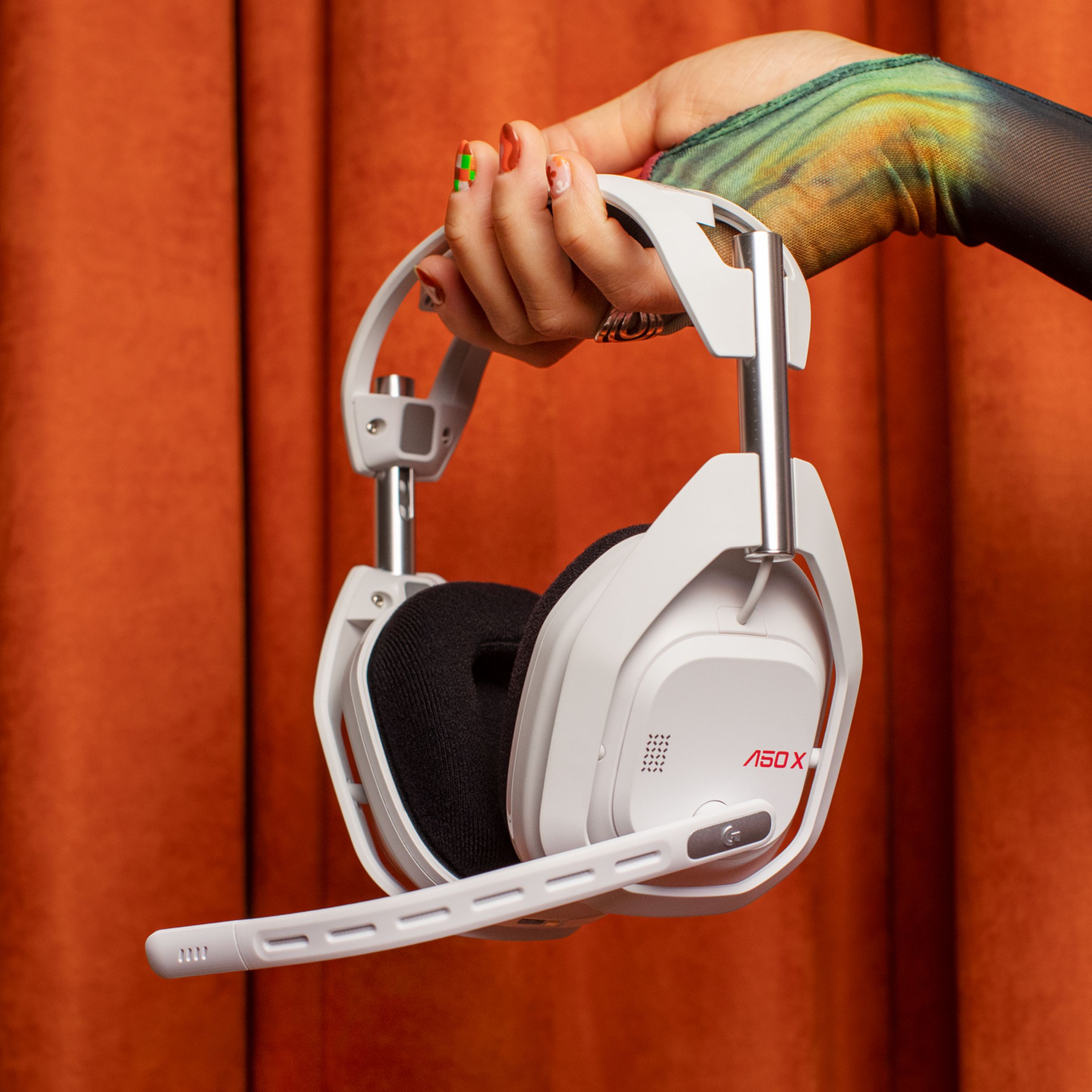 A person’s hand holding the white Astro A50 X gaming headset in front of a fabric backdrop.
