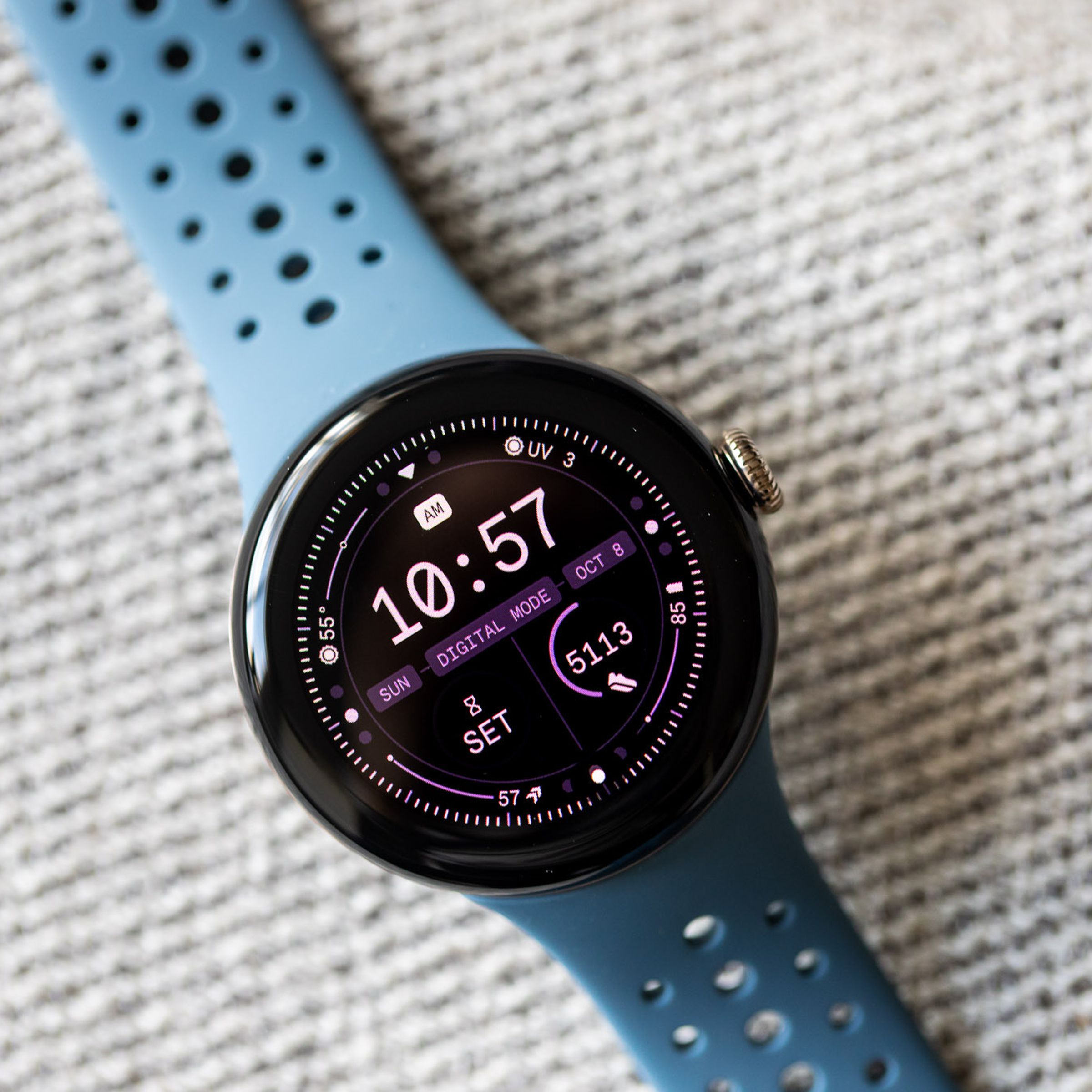 The second-generation Google Pixel Watch with a blue band resting on a surface with its display on. 