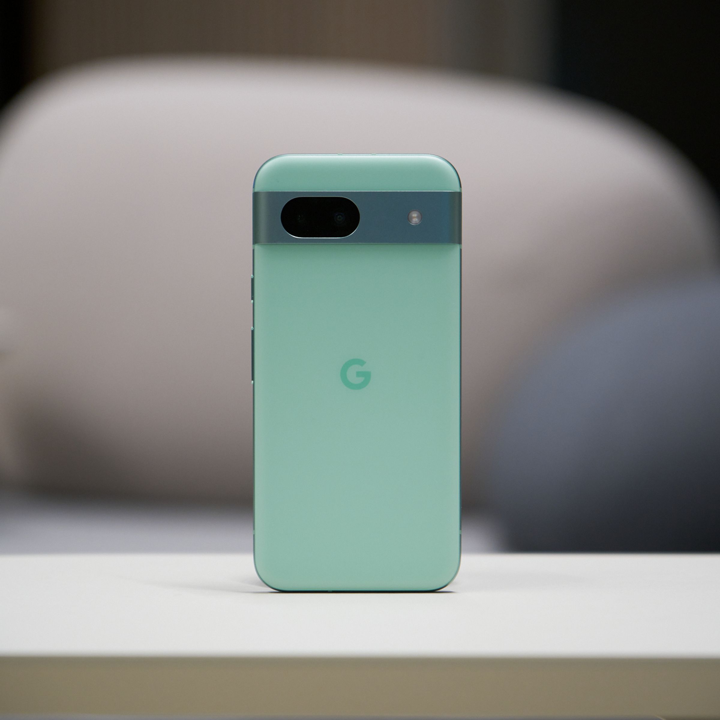 An image of an aloe-colored Google Pixel 8A placed upright in the center of a table.