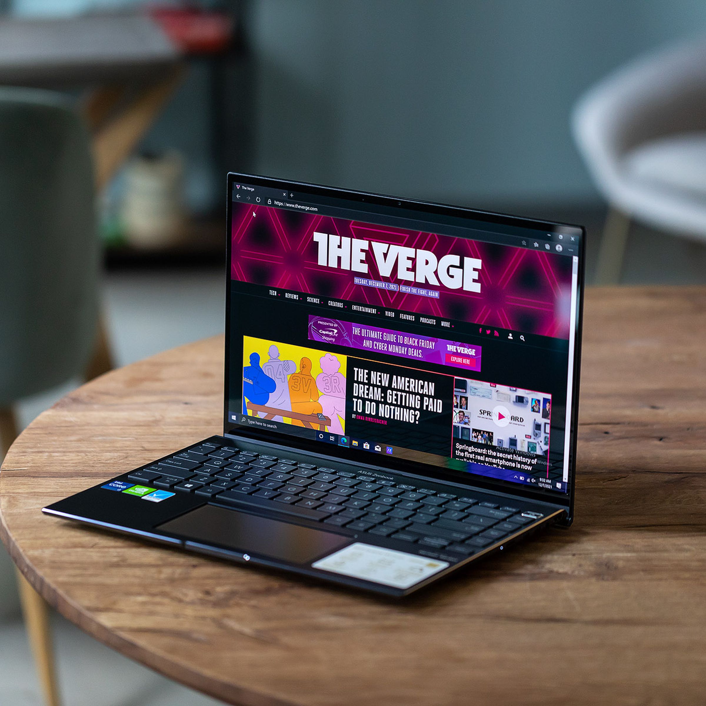 The Zenbook 14X OLED on a wooden table angled to the left. The screen displays The Verge homepage.
