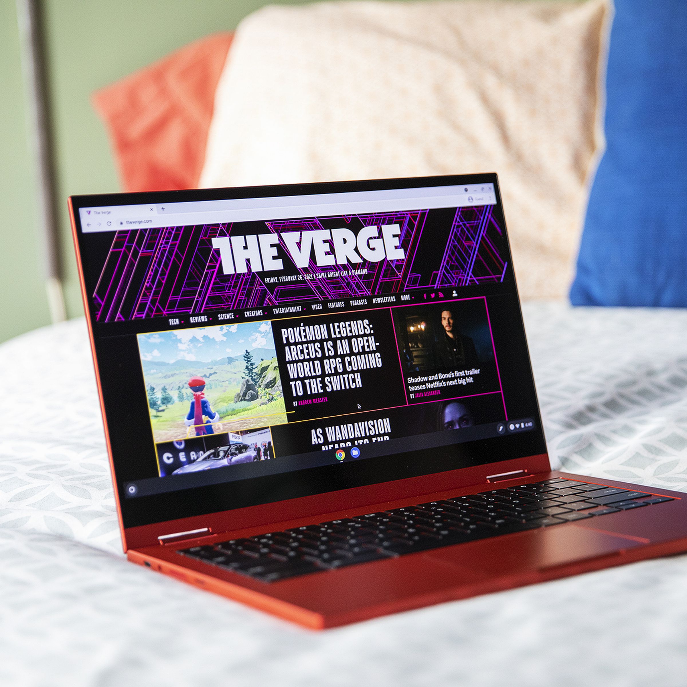The Samsung Galaxy Chromebook open, angled slightly to the right on the corner of a bed. The screen displays The Verge homepage.