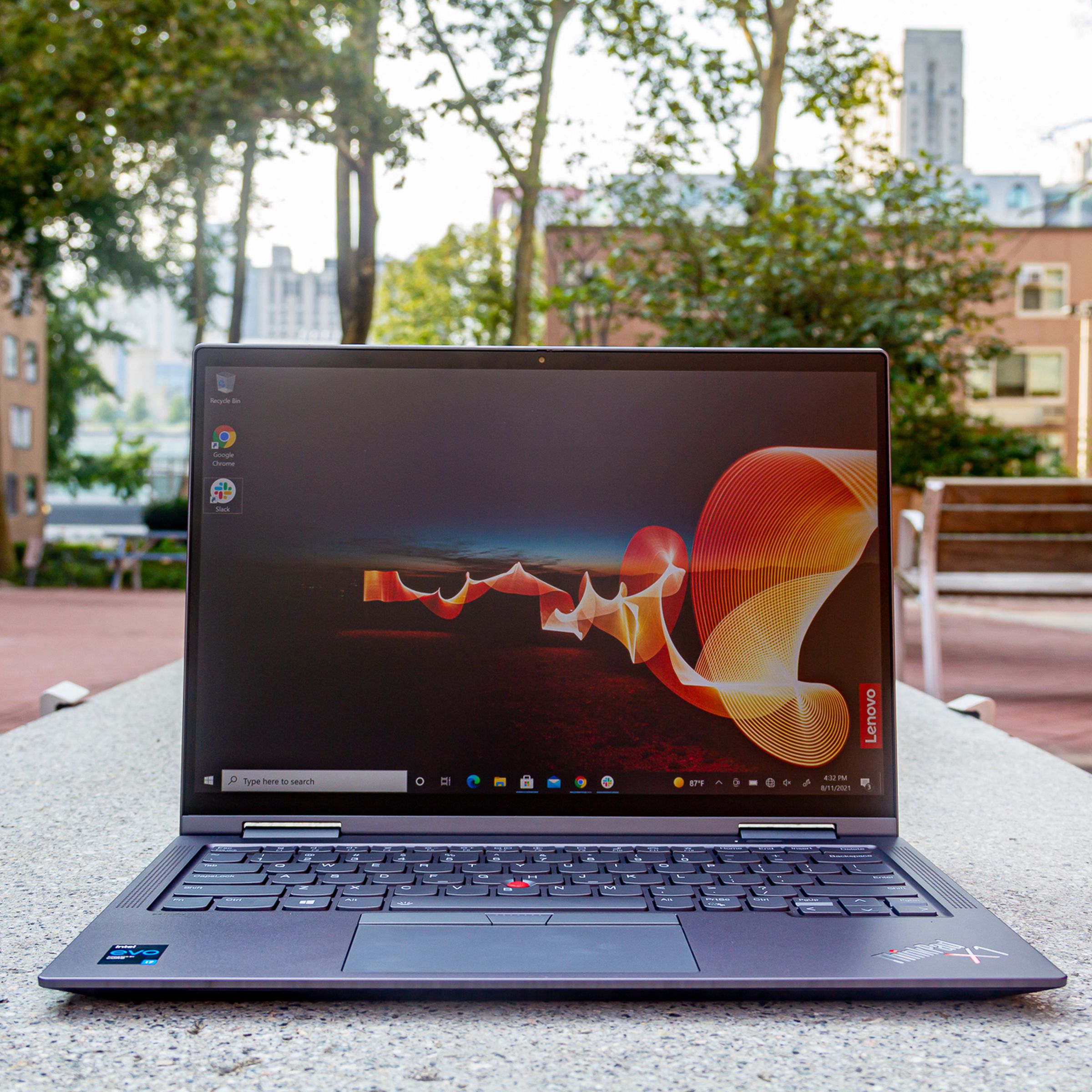 The ThinkPad X1 Yoga Gen 6 on a bench in an outdoor park, open. The screen displays an outdoor night scene with an orange banner rippling across it and the Lenovo banner on the bottom right side.