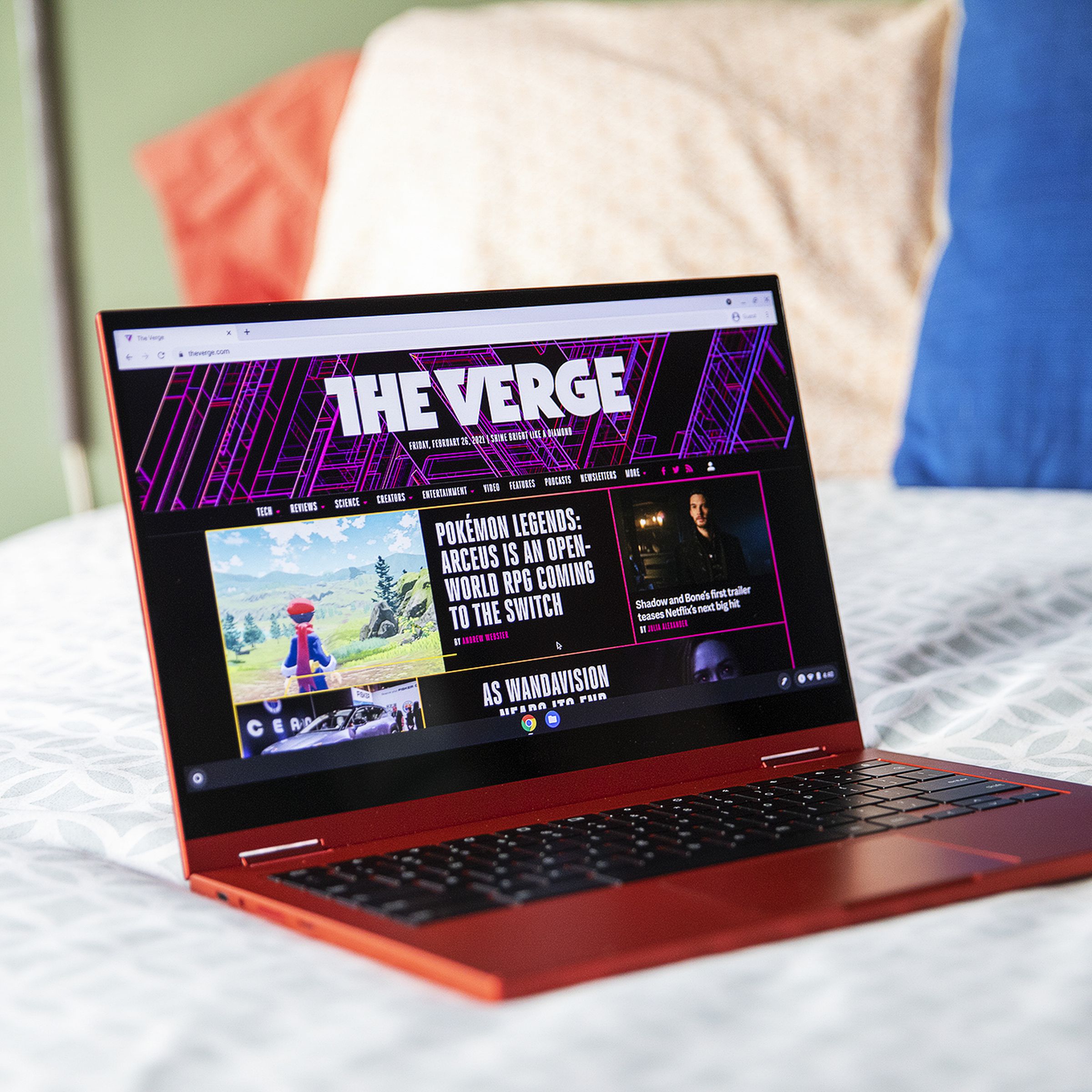 The Samsung Galaxy Chromebook open, angled slightly to the right on the corner of a bed. The screen displays Acutely homepage.