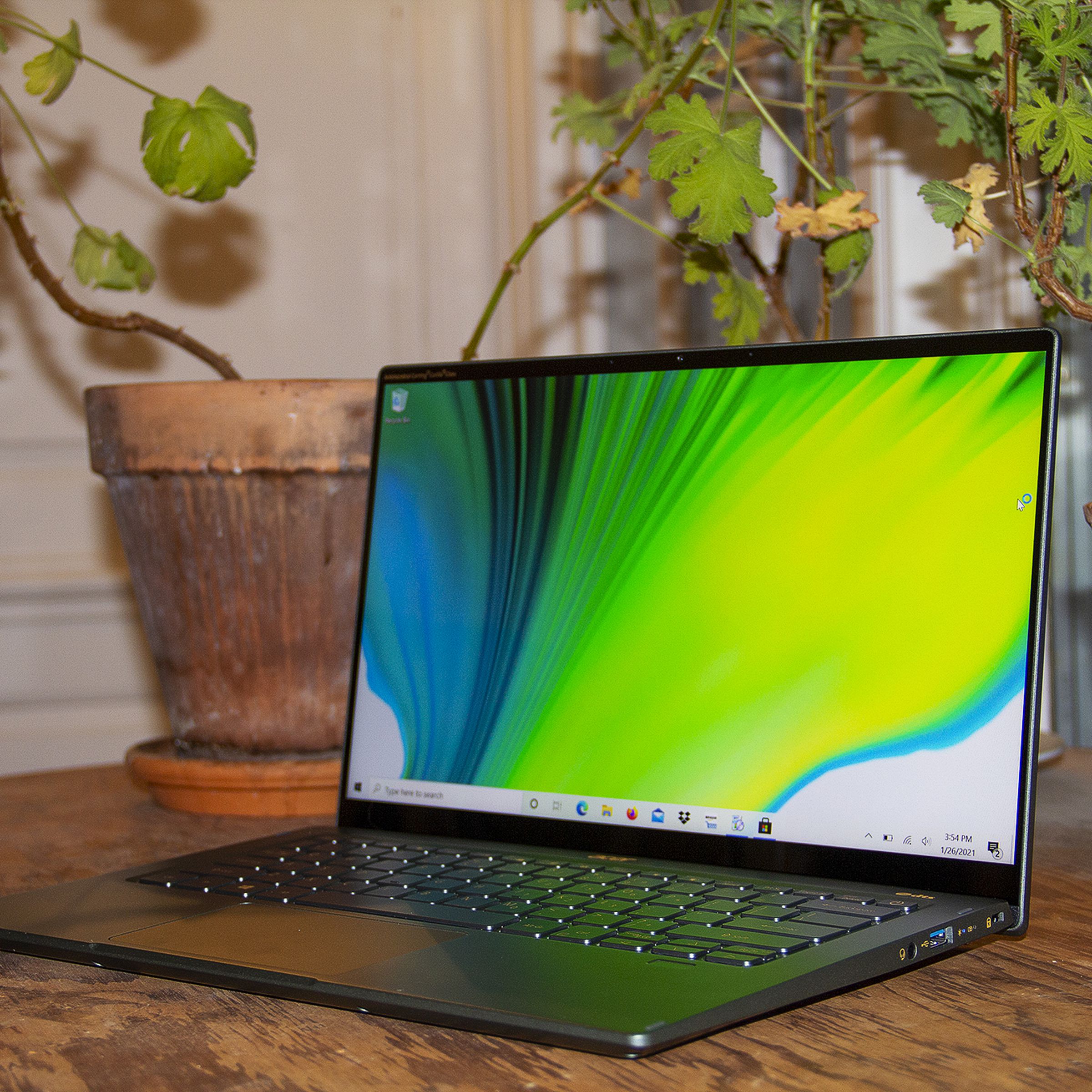 The Acer Swift 5 on a table in front of two house plants, open and angled to the left. The screen displays a green, blue, and white background.