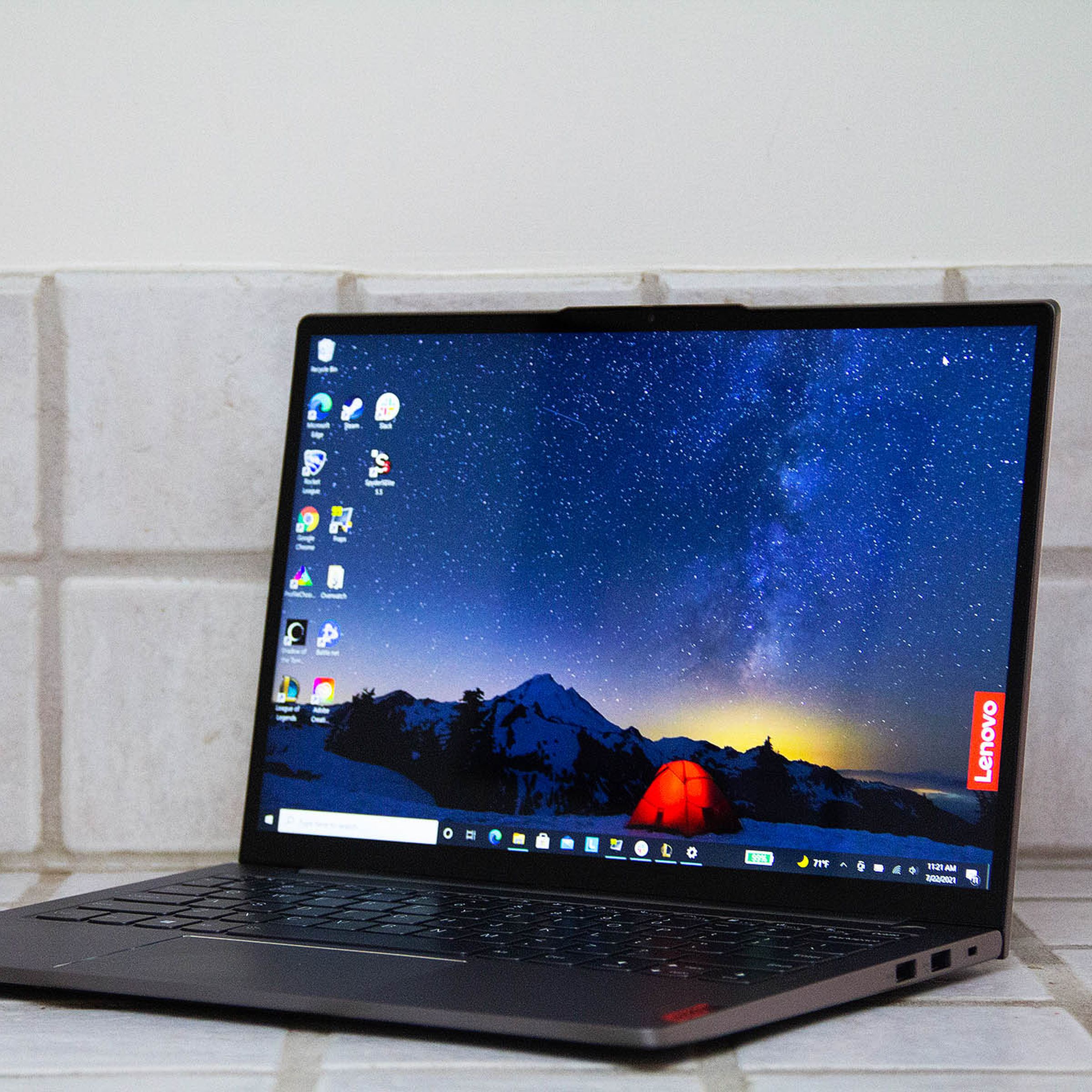 The Lenovo ThinkBook 13s on a white tiled counter, open, angled to the left. The screen displays a night mountain scene with a red tent in the center and the red Lenovo banner on the right side.