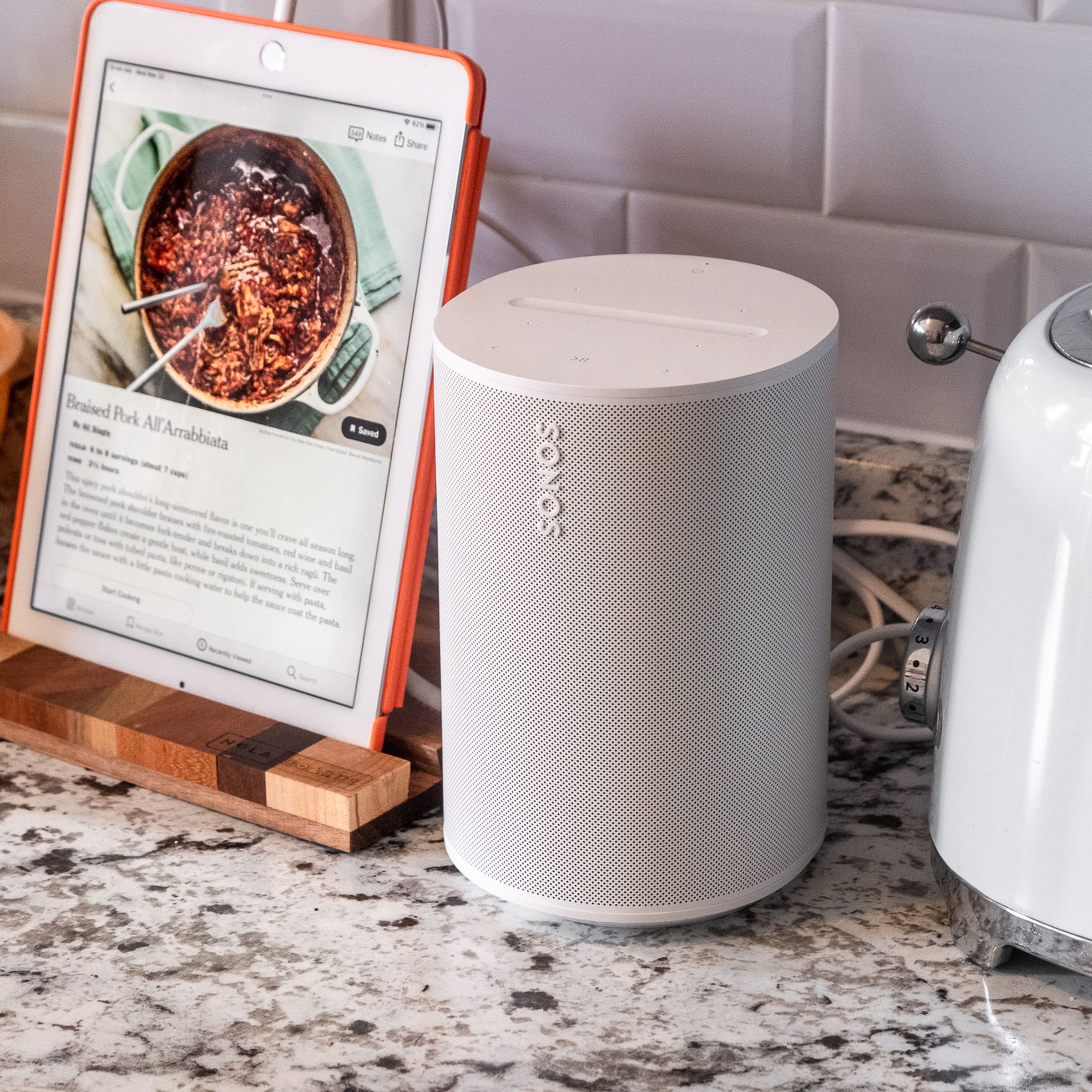 A photo of the Sonos Era 100 on a kitchen counter beside a toaster and iPad.