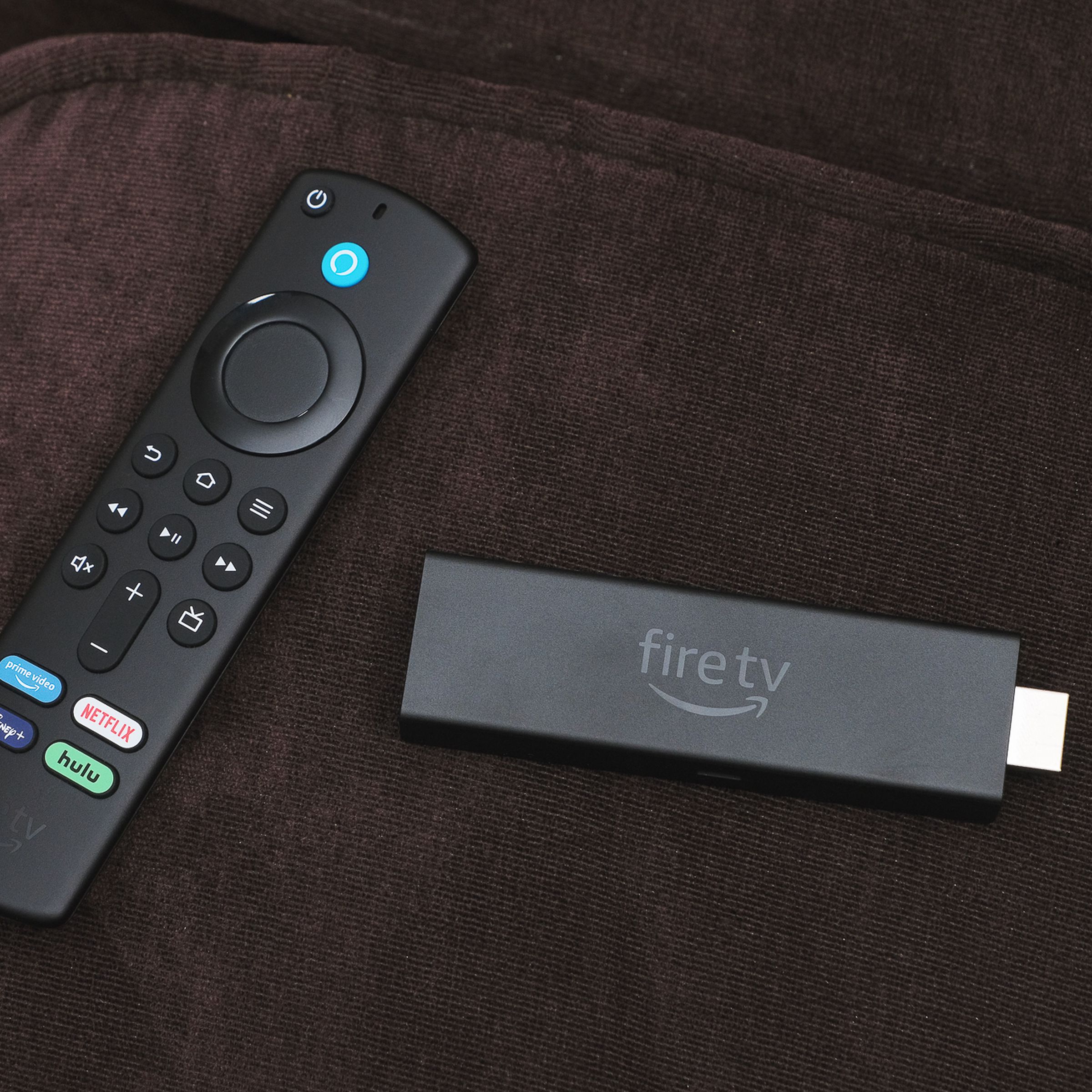 A close-up of the Amazon Fire TV Stick 4K Max streaming dongle beside its remote control on a brown couch.