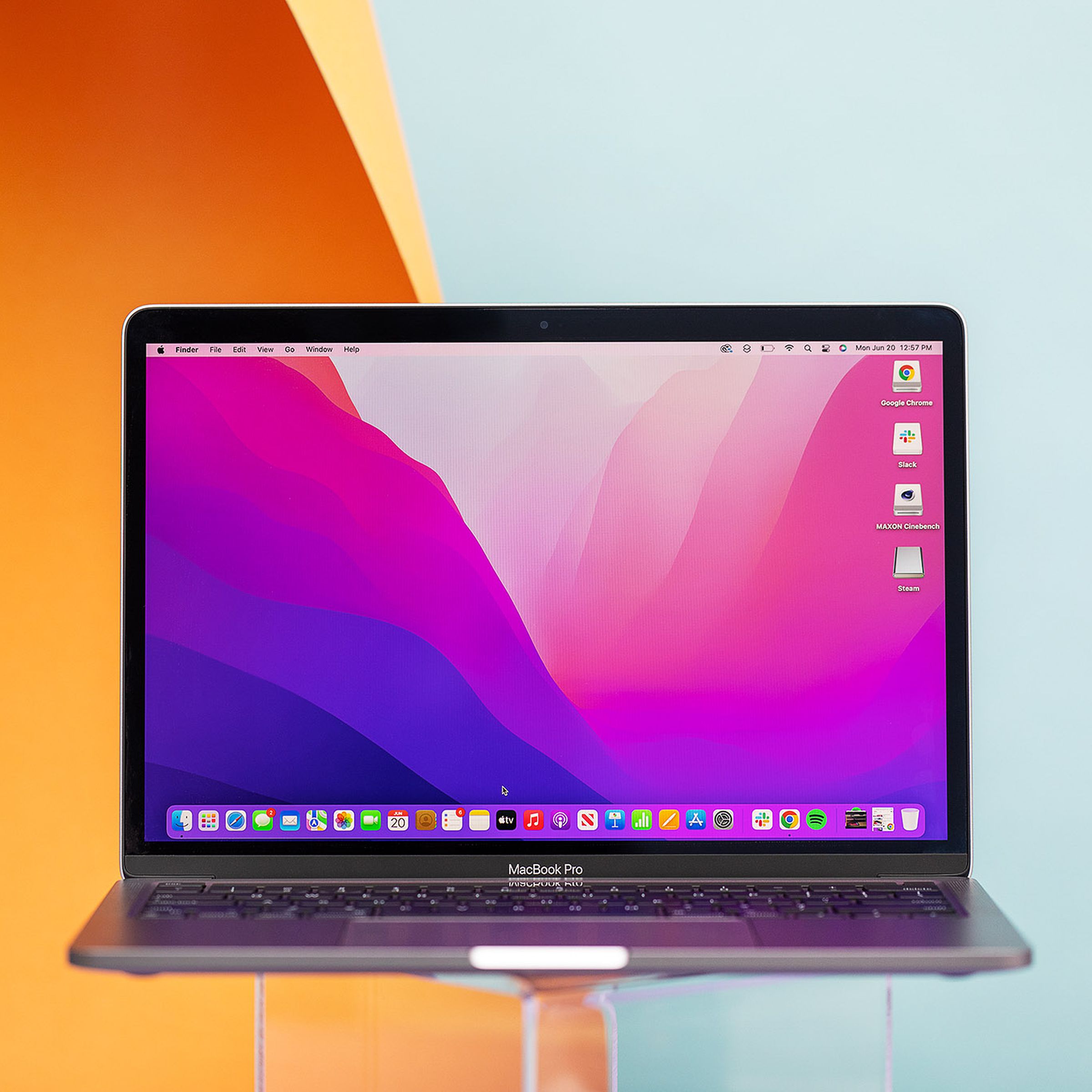 MacBook Pro 13 2022, şeffaf bir stand üzerinde açılır. Ekranda pembe ve mor bir masaüstü arka planı görüntülenir.