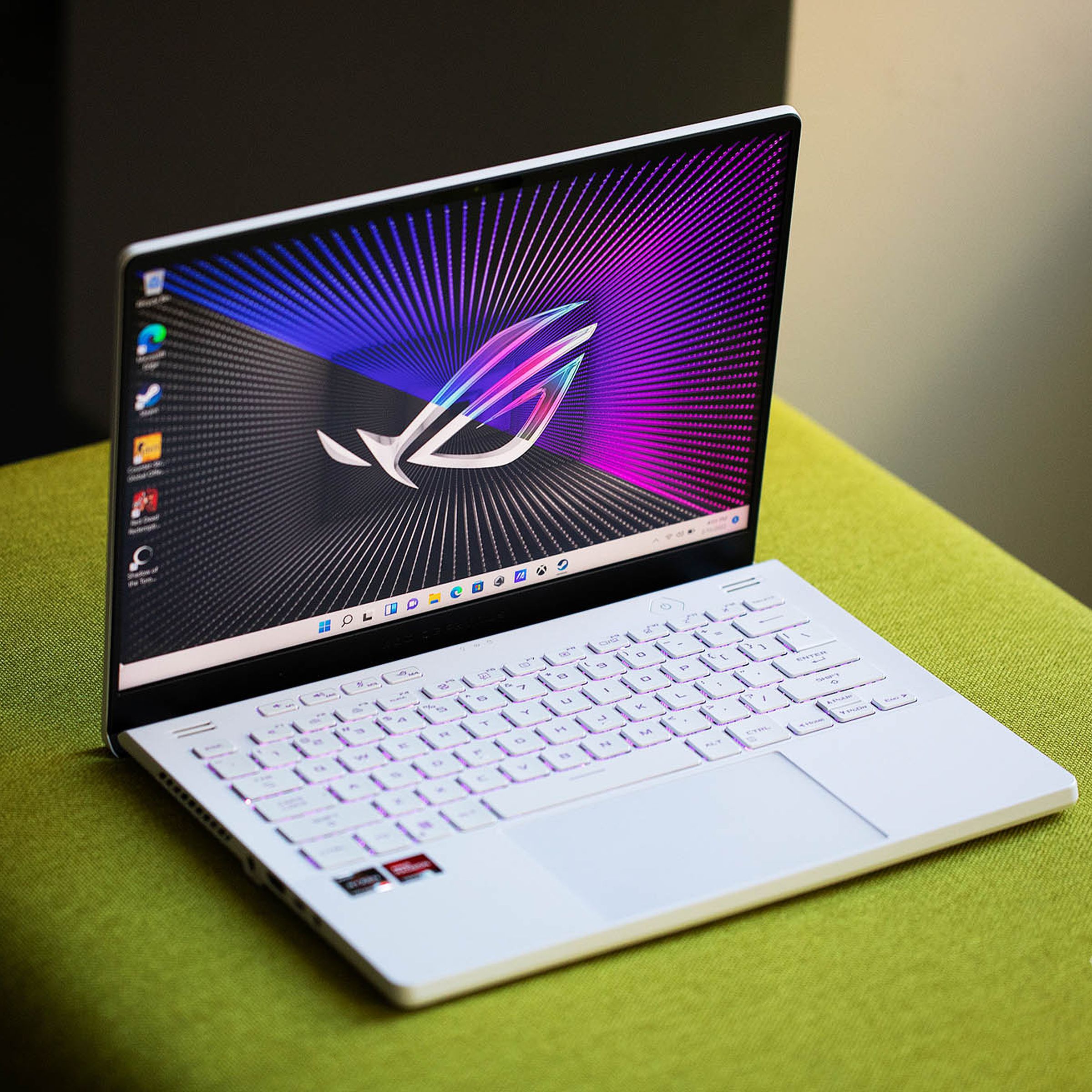 A white Asus thin gaming laptop on a green fabric surface, displaying a big Asus logo on a predominantly purple desktop background