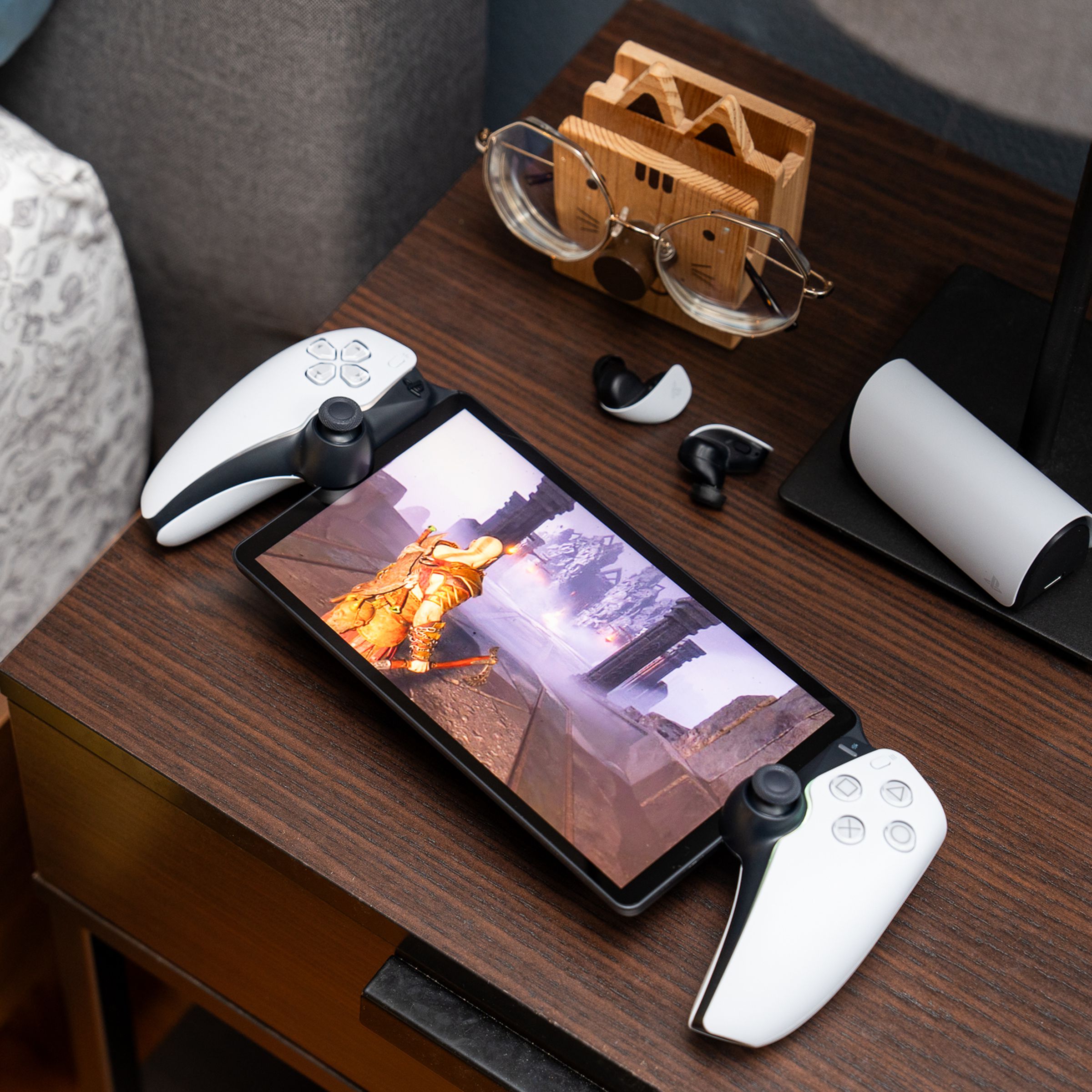 The PlayStation Portal sitting on a bedside table with a pair of earbuds. The handheld gaming device is streaming God of War: Ragnarök off a PlayStation 5.