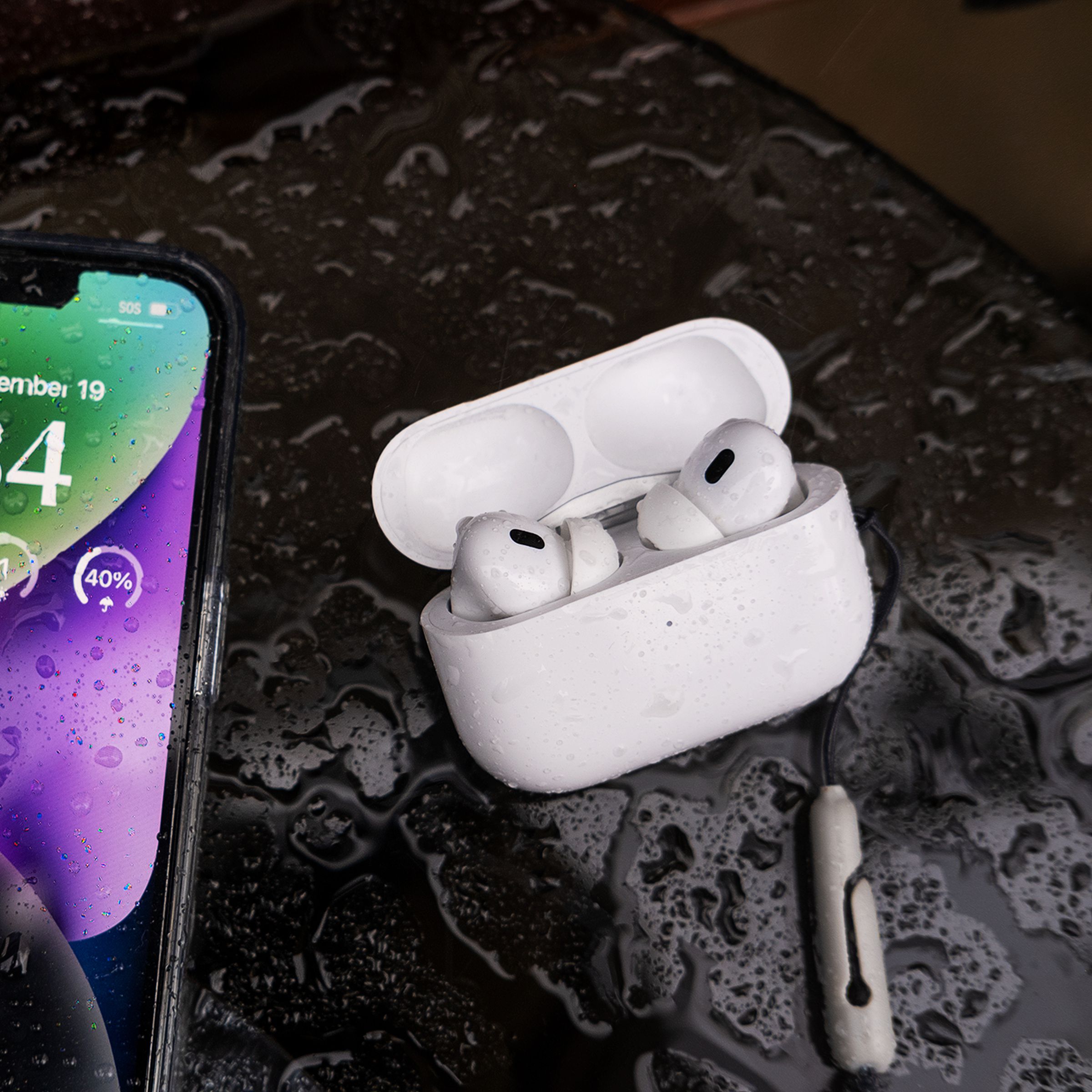 A photo of the iPhone 14 next to Apple’s second-generation AirPods Pro. Both devices are wet with visible raindrops on them.
