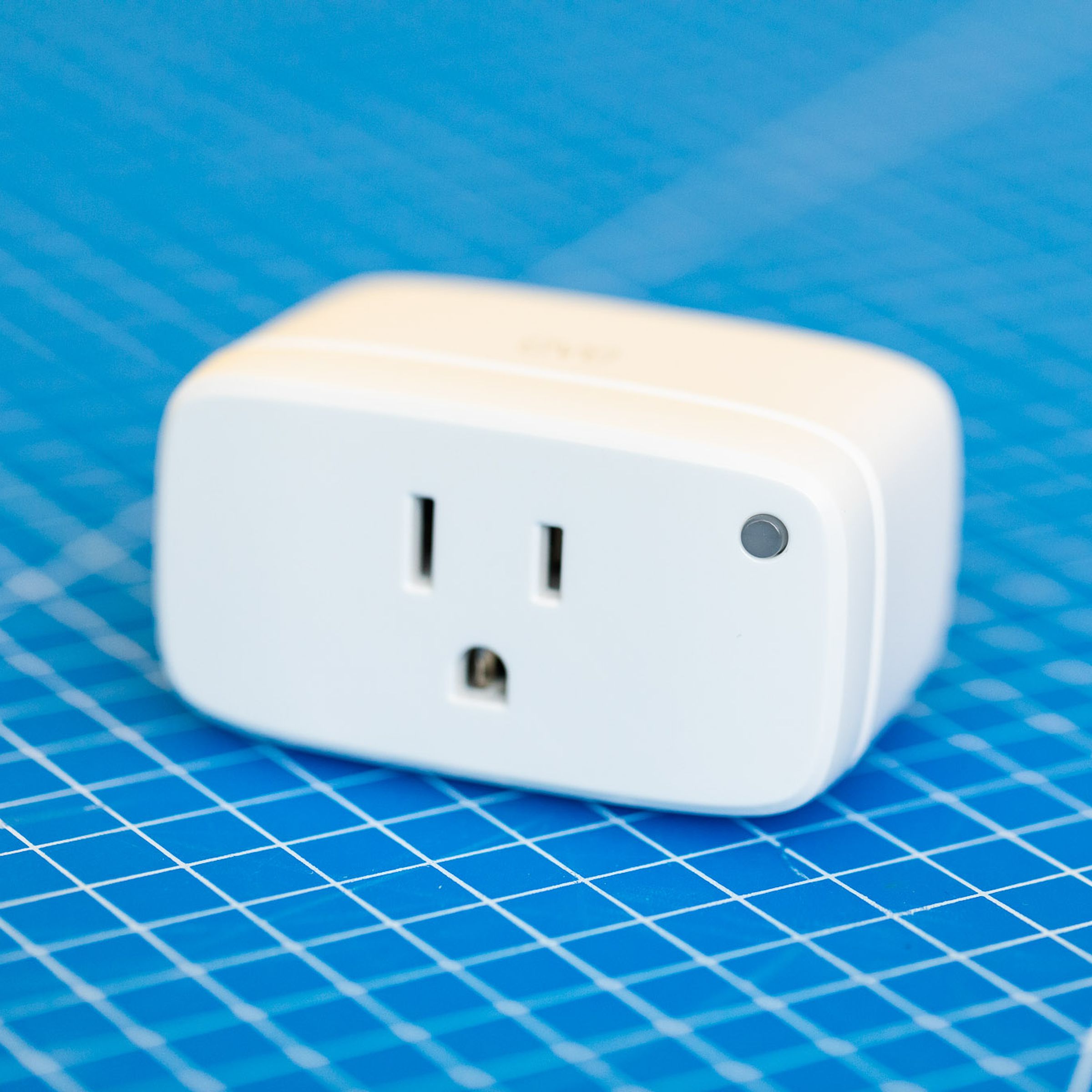 Eve Energy smart plug. A white rectangular prism with the broad face front. The corners and edges are rounded, there is a three-prong outlet on the front and a green LED on the upper-right corner of the front face. It sits on a blue background with a grid of white lines.