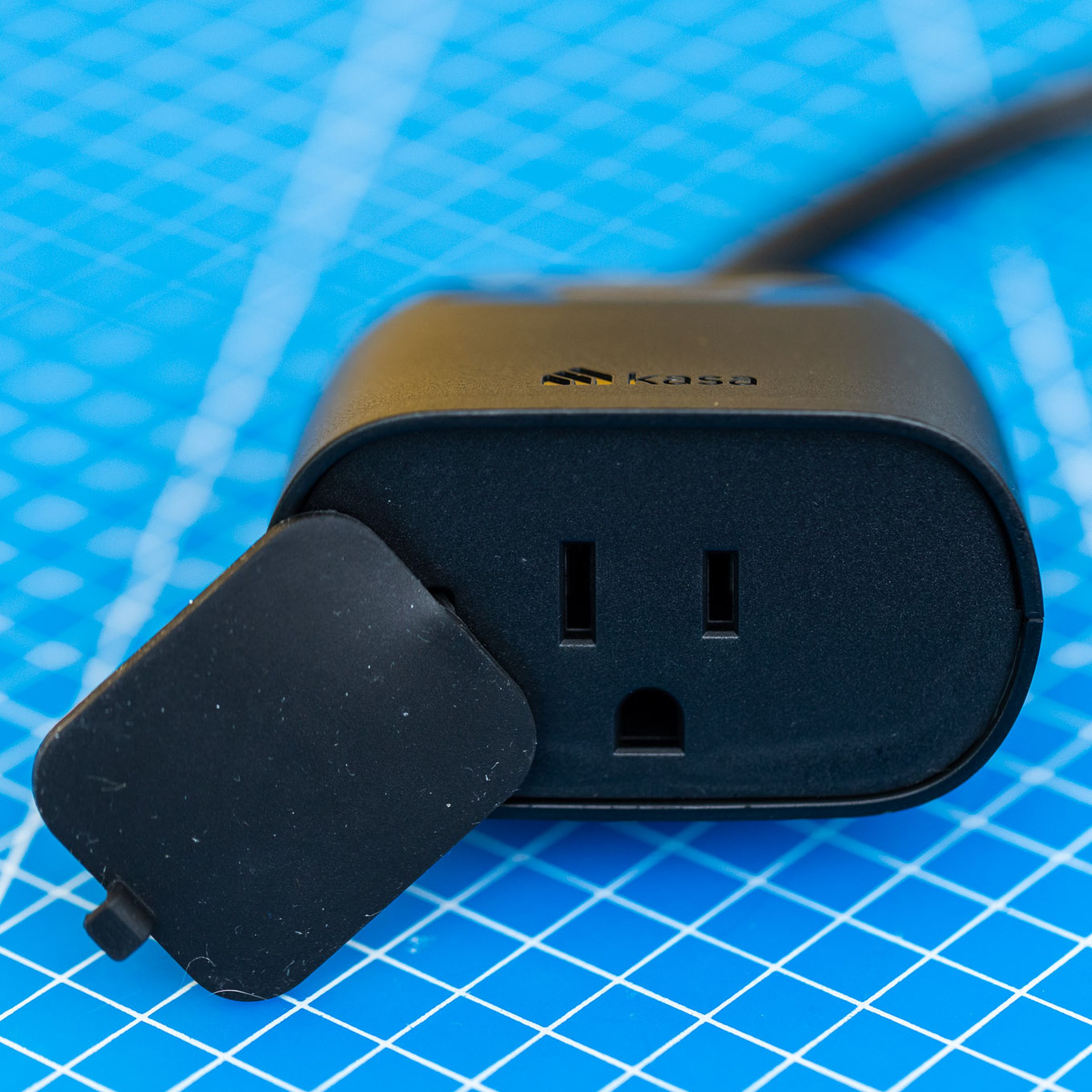 A black, two-outlet outdoor smart plug. It has a short cable leading to its own power plug, and a flap to protect the outlets from moisture. It is on a blue background with a grid of white lines.