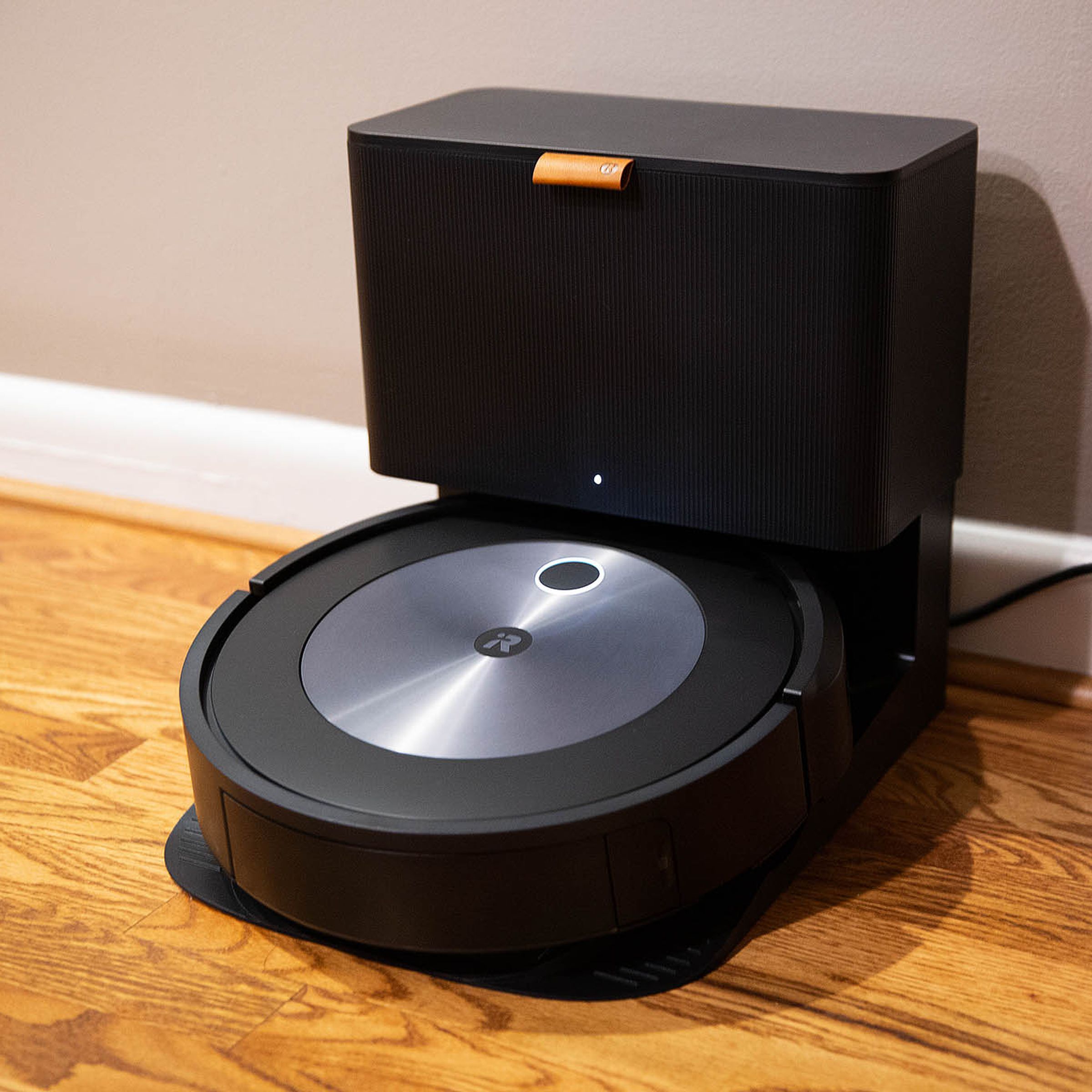 Image of a black-and-silver disc-shaped robot vacuum docked into a black auto-empty dock. The dock has an orange tag on the front and a small white status LED on the lower front face above where the vacuum sits at rest.