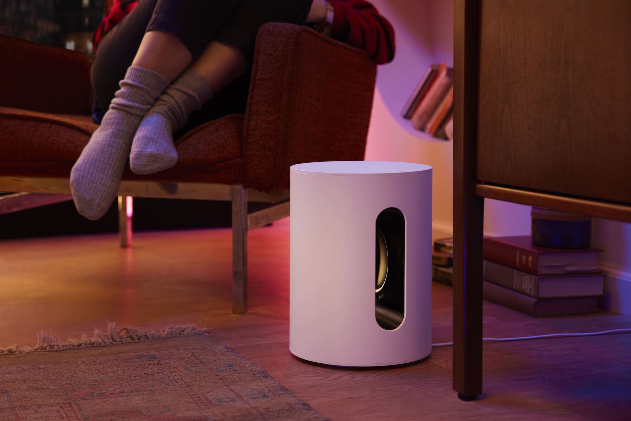 A Sonos Sub Mini subwoofer shown on the floor with someone sitting nearby.