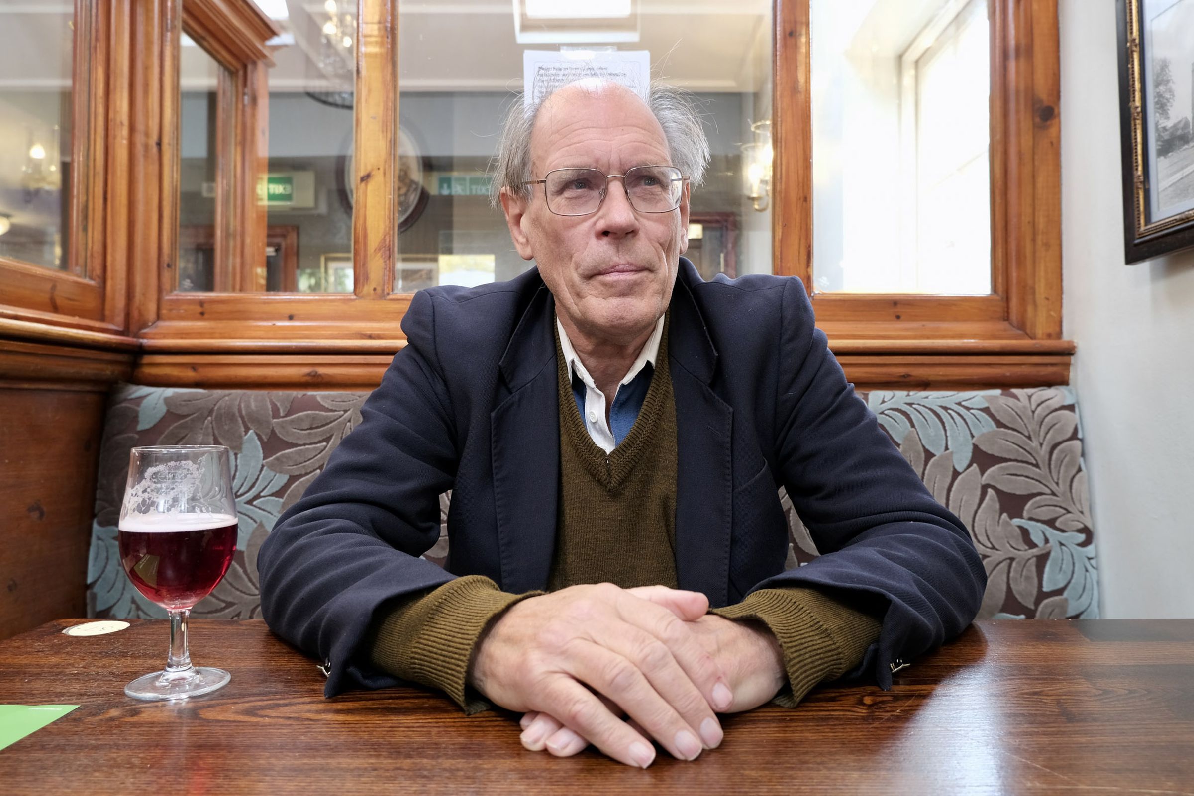 A photo of a man sitting in a pub. He’s balding, wearing spectacles, and suit jacket over a V-neck jumper. His hands are folded in front of him on the table next to a half-drunk pint of beer. 