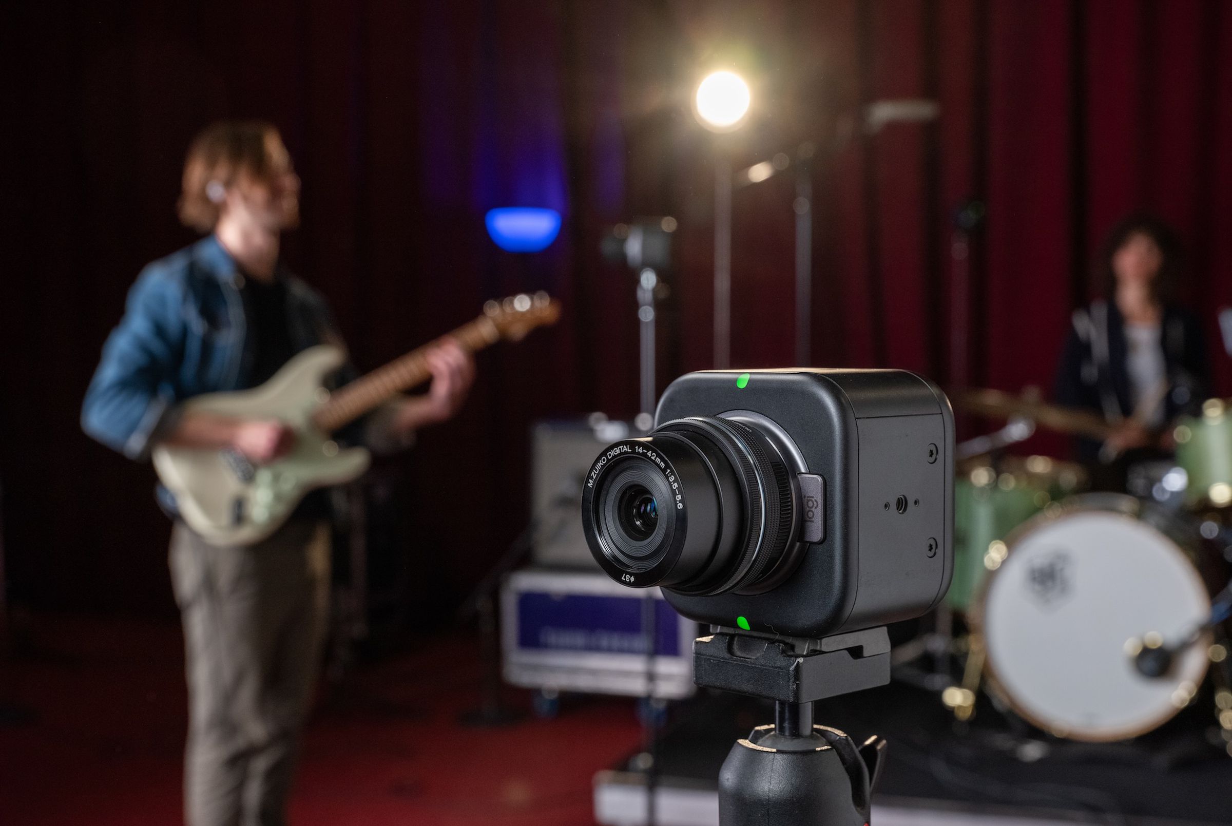 An image of Logitech’s Mevo Core camera in the foreground with a rock band playing in the background.