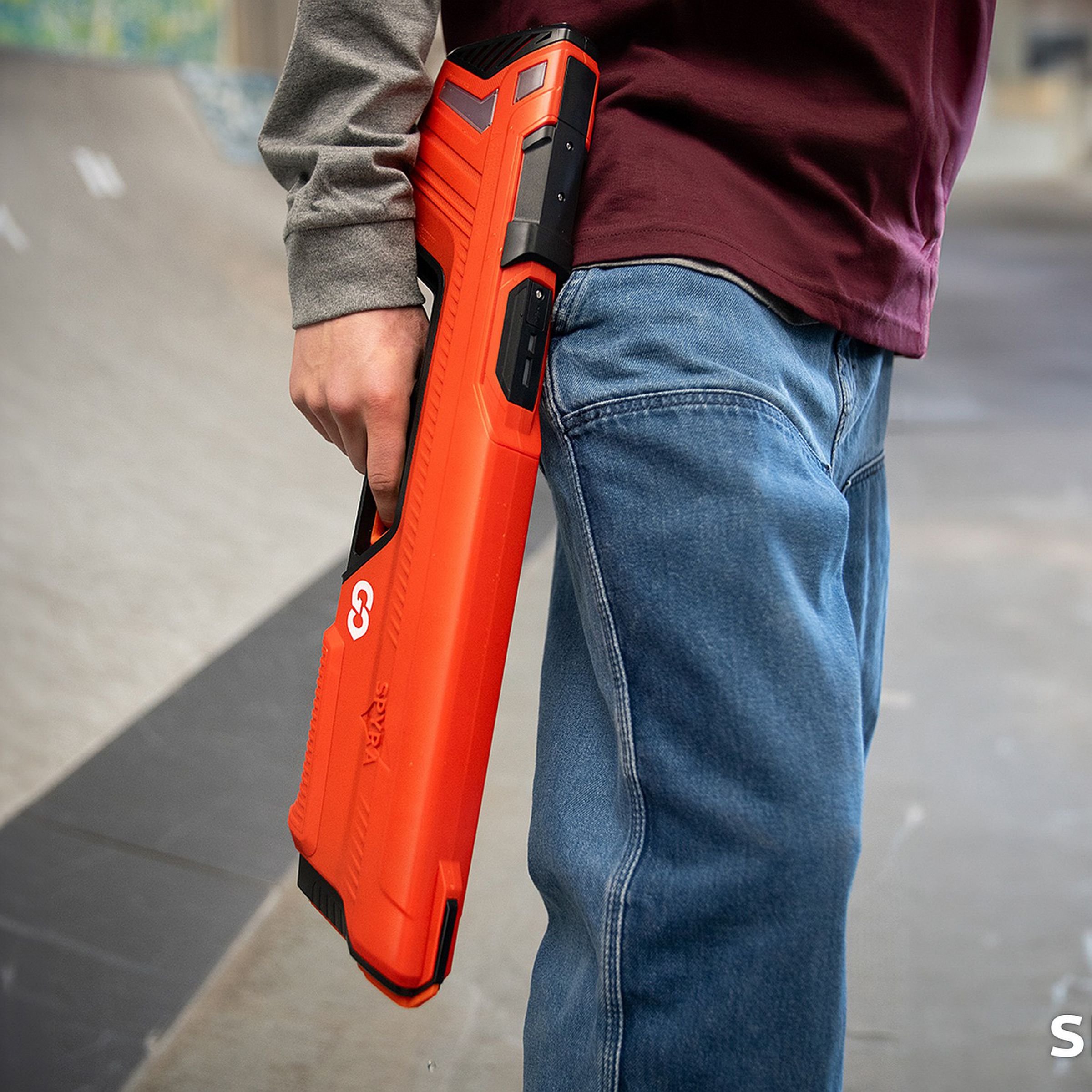 A red, raking toy rifle held by a teen’s arm, pointed down along the same line as their blue jeans