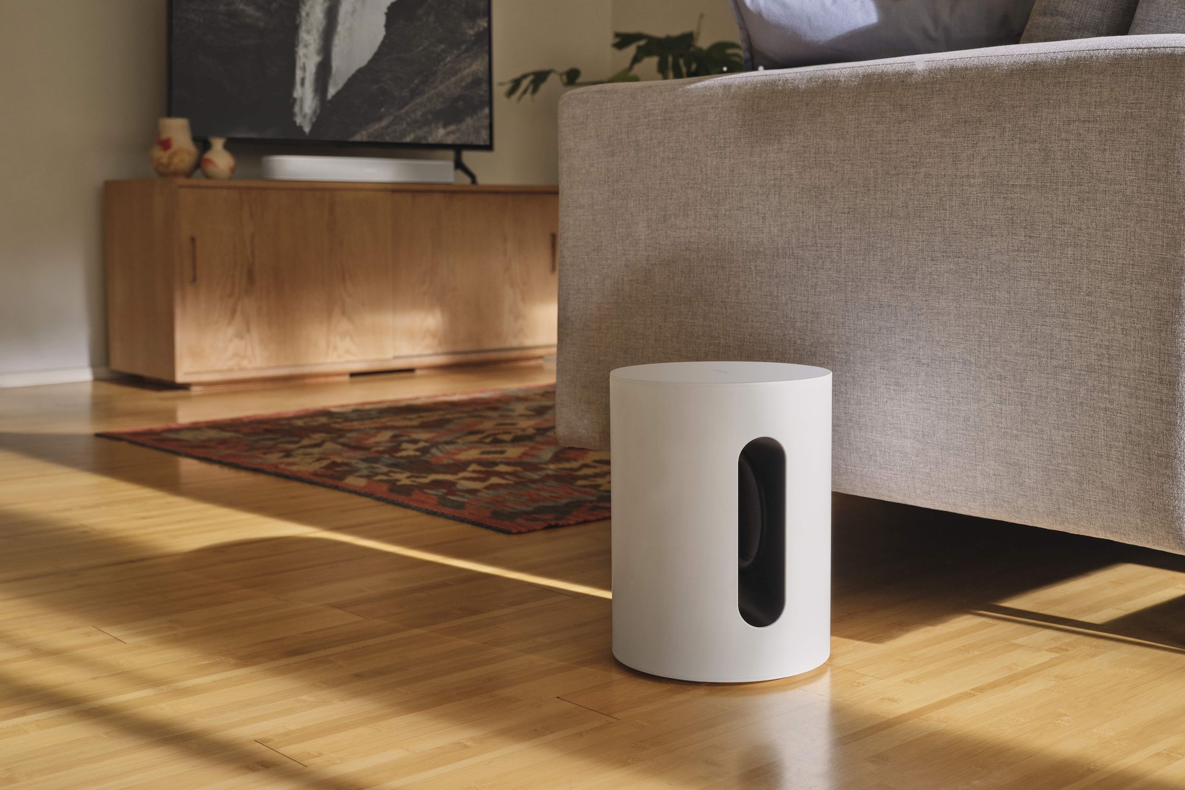 A Sonos Sub Mini subwoofer pictured next to a couch, with a television in the background.