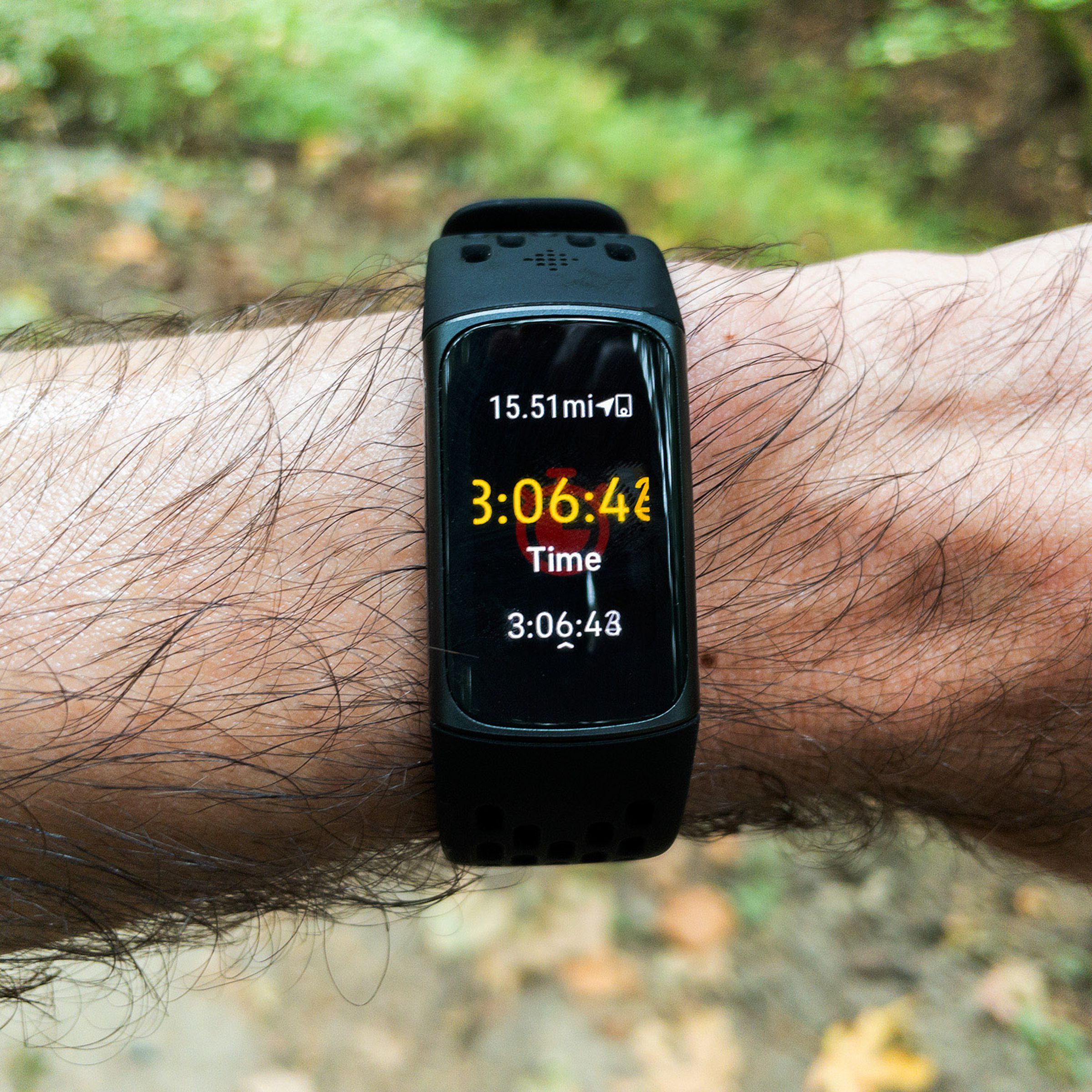 Close-up of a Fitbit Charge 5 worn on a person’s left wrist. There appears to be a forest path in the background. The person seems to be on a long run; the display reads “15.51 mi” with an elapsed time of 3 hours, 6 minutes, and 41 seconds. 