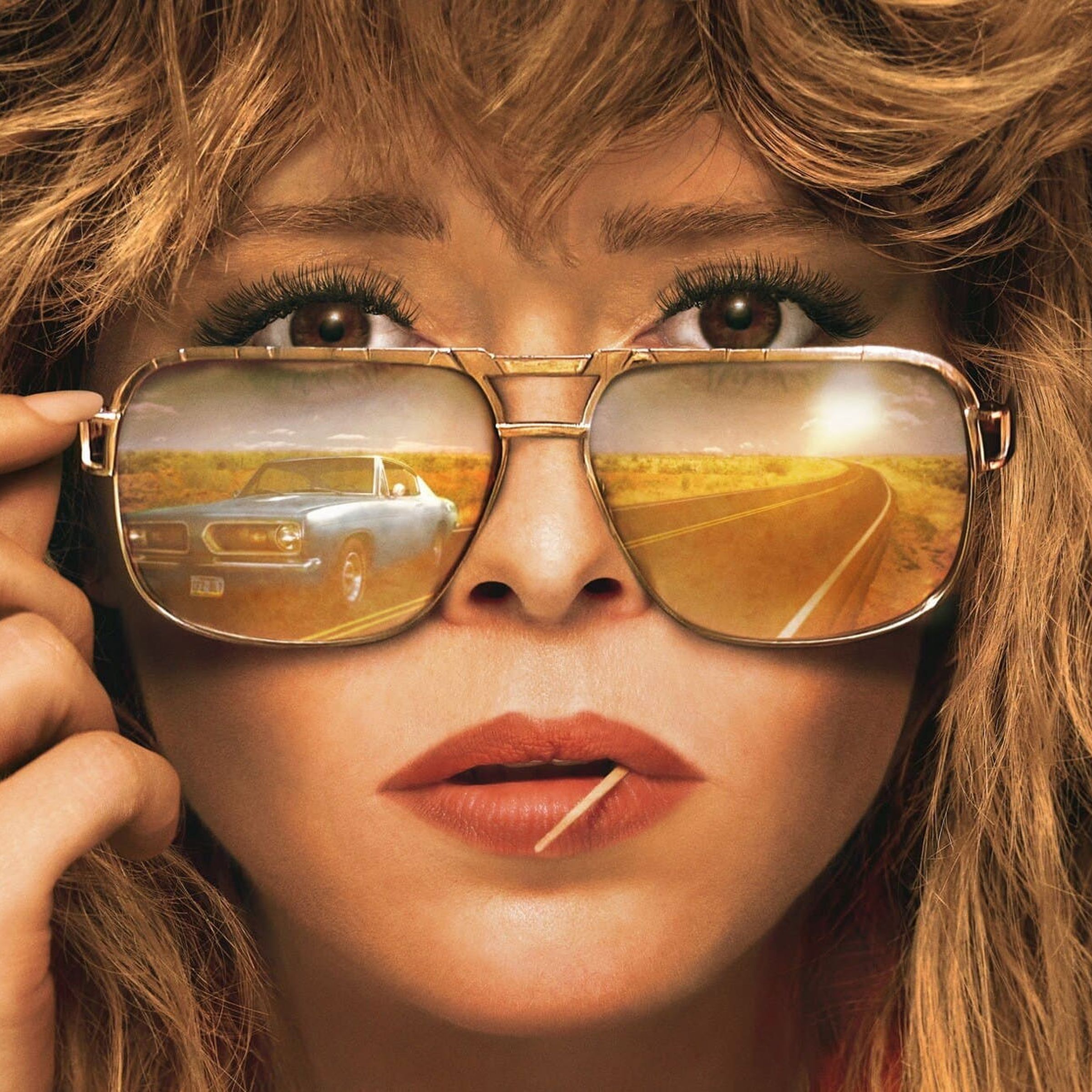 A blonde woman with layered hair, wearing a pair of aviators in which her blue car and the highway are being reflected.