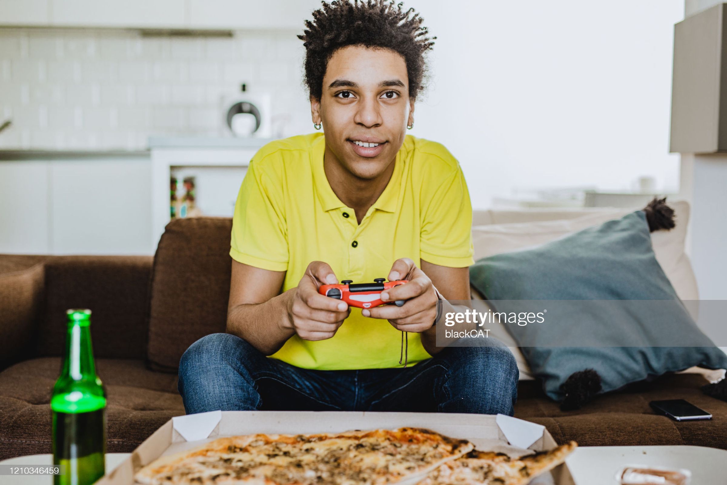 “Young African American man eating pizza, drinking beer and playing video games.”
