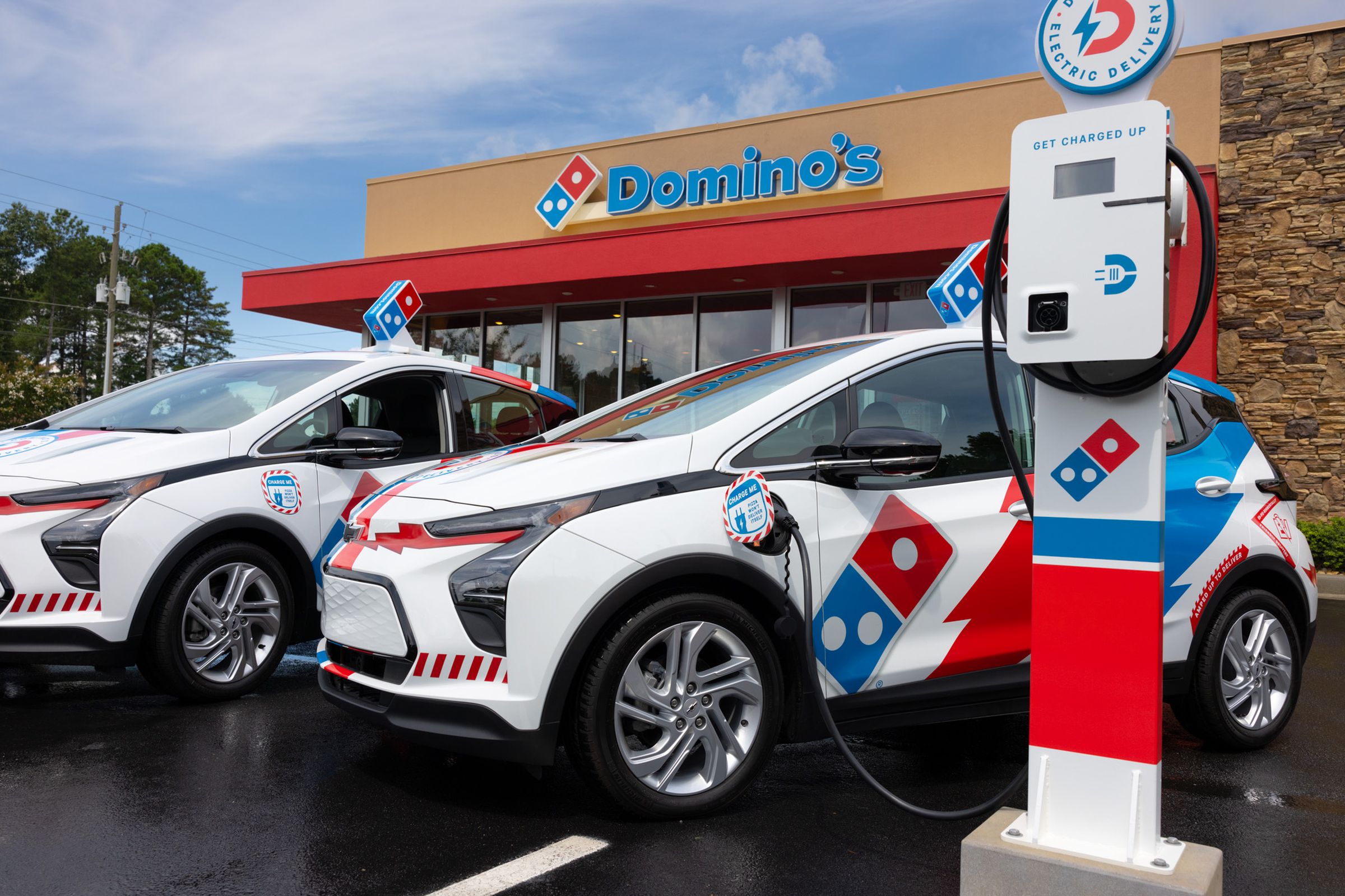 Two chevy bolt ev cars, wrapped in domino’s artwork are parked in front of a domino’s pizza store with one of the cars hooked up to a charger.