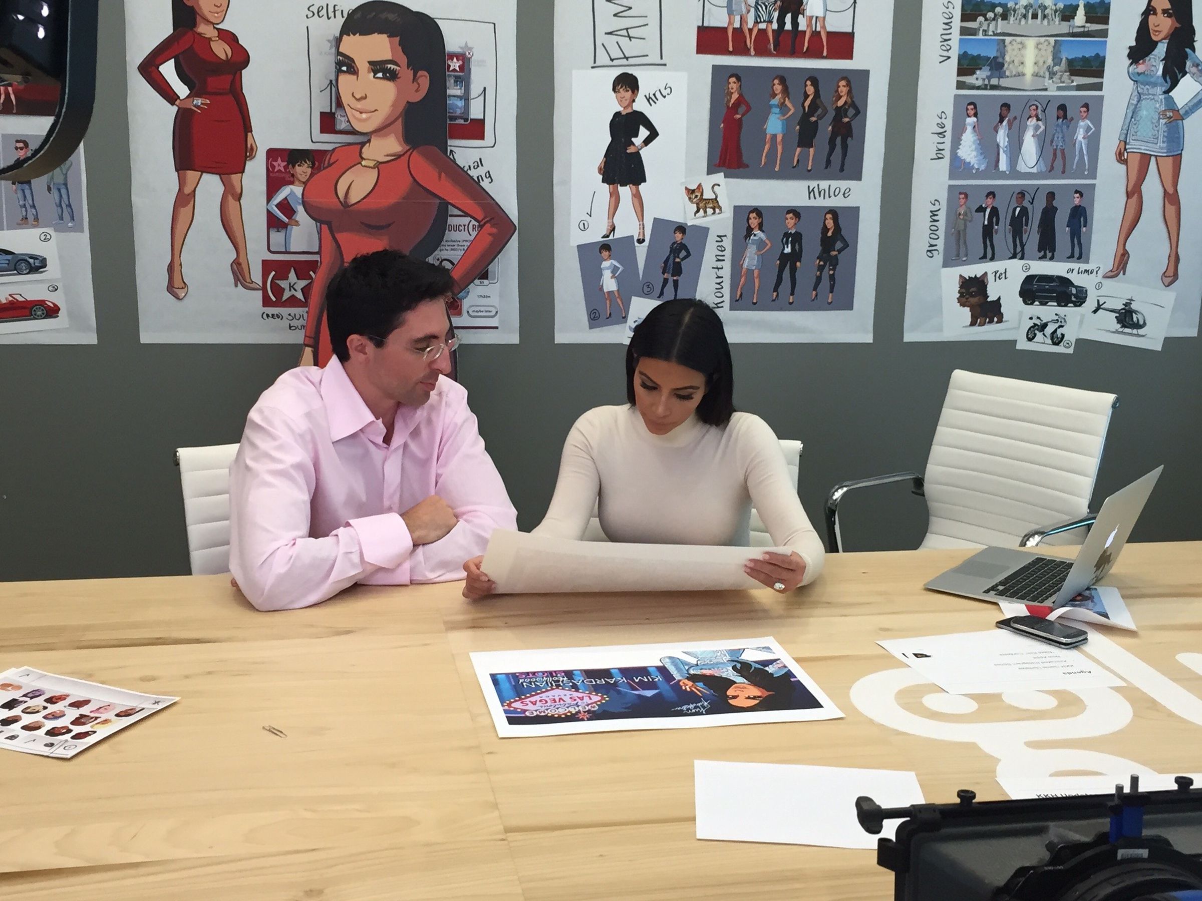 Kim Kardashian at a desk in the Glu Mobile offices looking over designs for the game, with concept art on the wall behind her