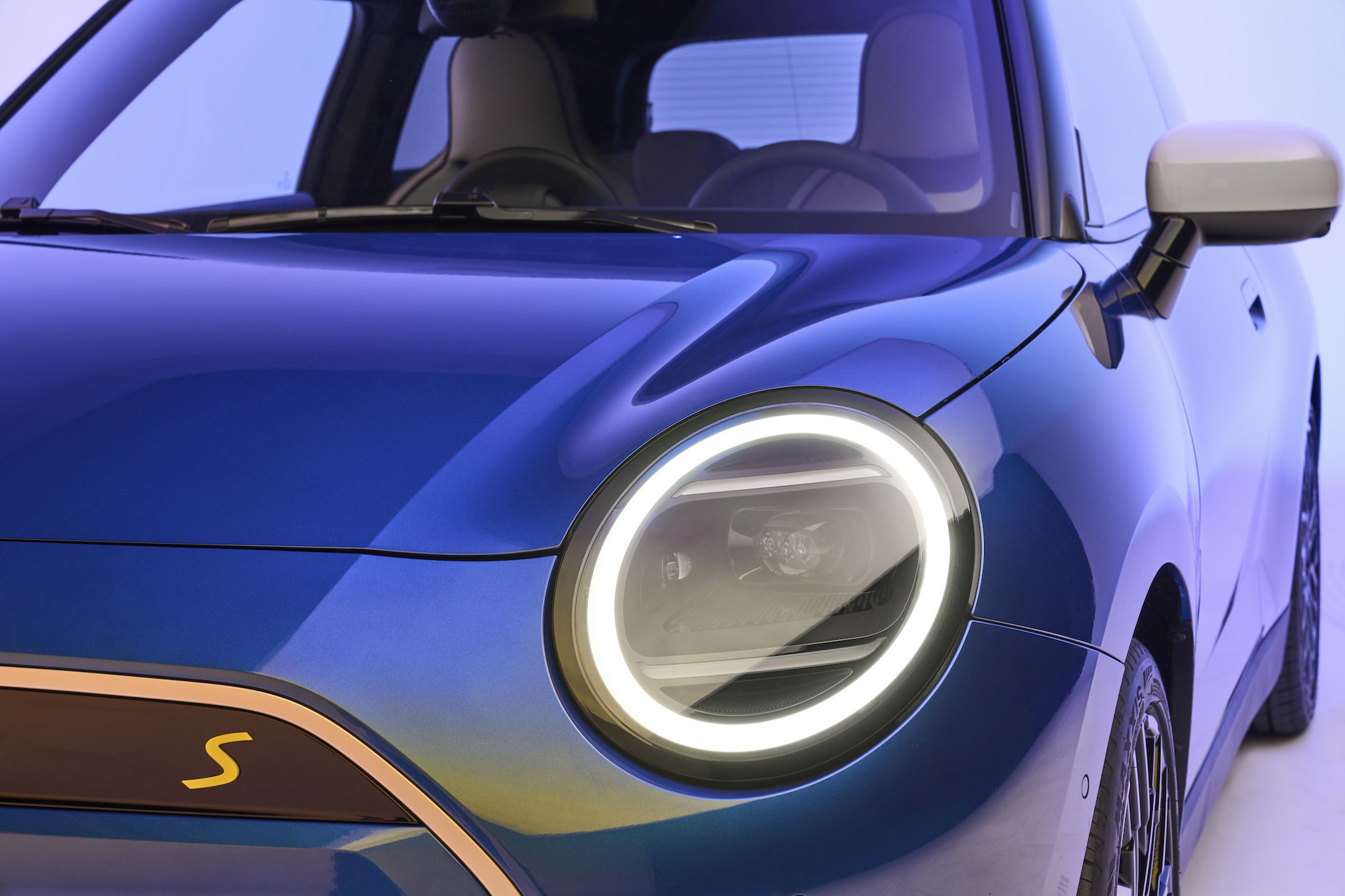 Close-up of the driver’s side corner of a blue Mini Cooper EV, showing the bright ring around the headlight.