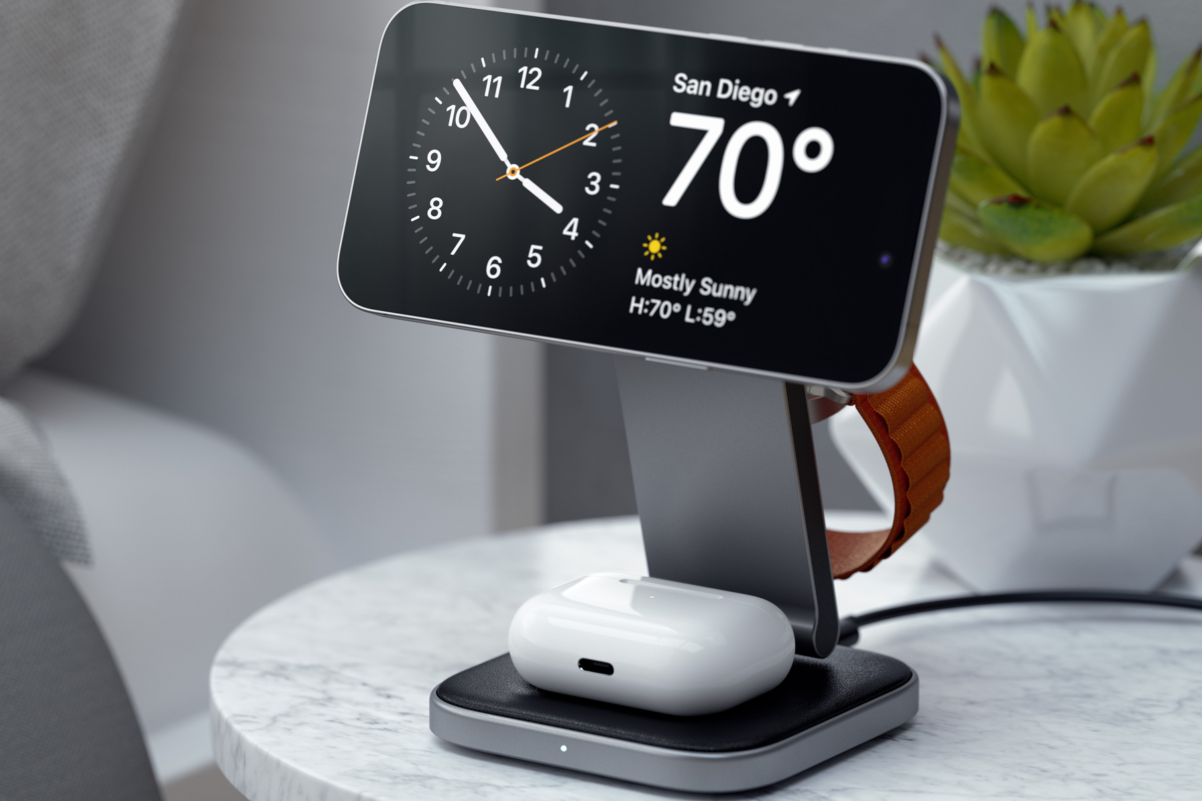A picture of an iPhone in landscape mode on a charging stand, suspended above an AirPods Pro case, with an Apple Watch Ultra band just visible suspended from the charging puck on the back of the stand.