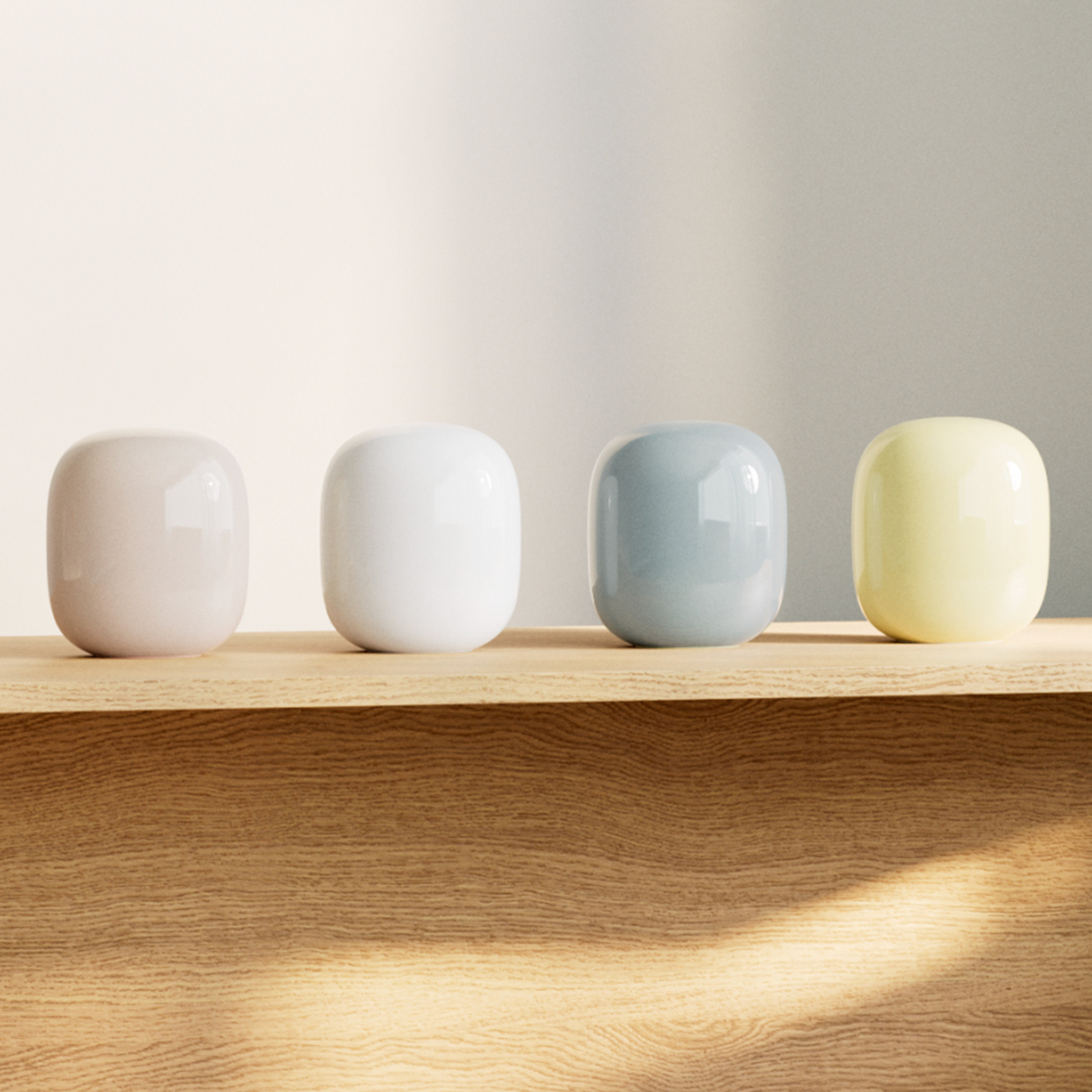 Photo of four Nest Wifi Pro routers on a wood table. One is pink, one is white, one is blue, and one is yellow. They are glossy and reflective rounded rectangles.