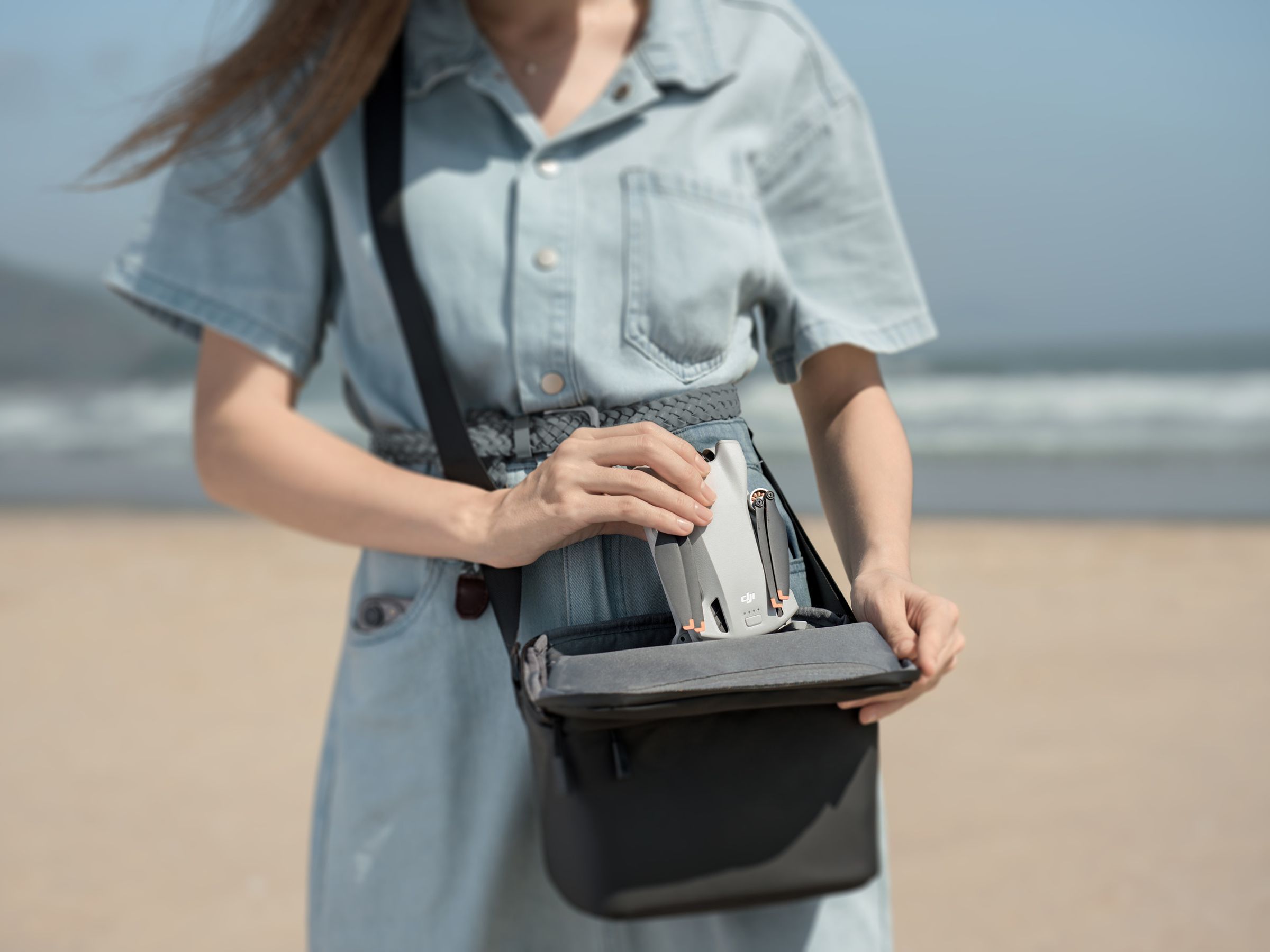 Une personne sur la plage, plaçant un drone DJI Mini 3 compacté dans un sac à main de taille moyenne.