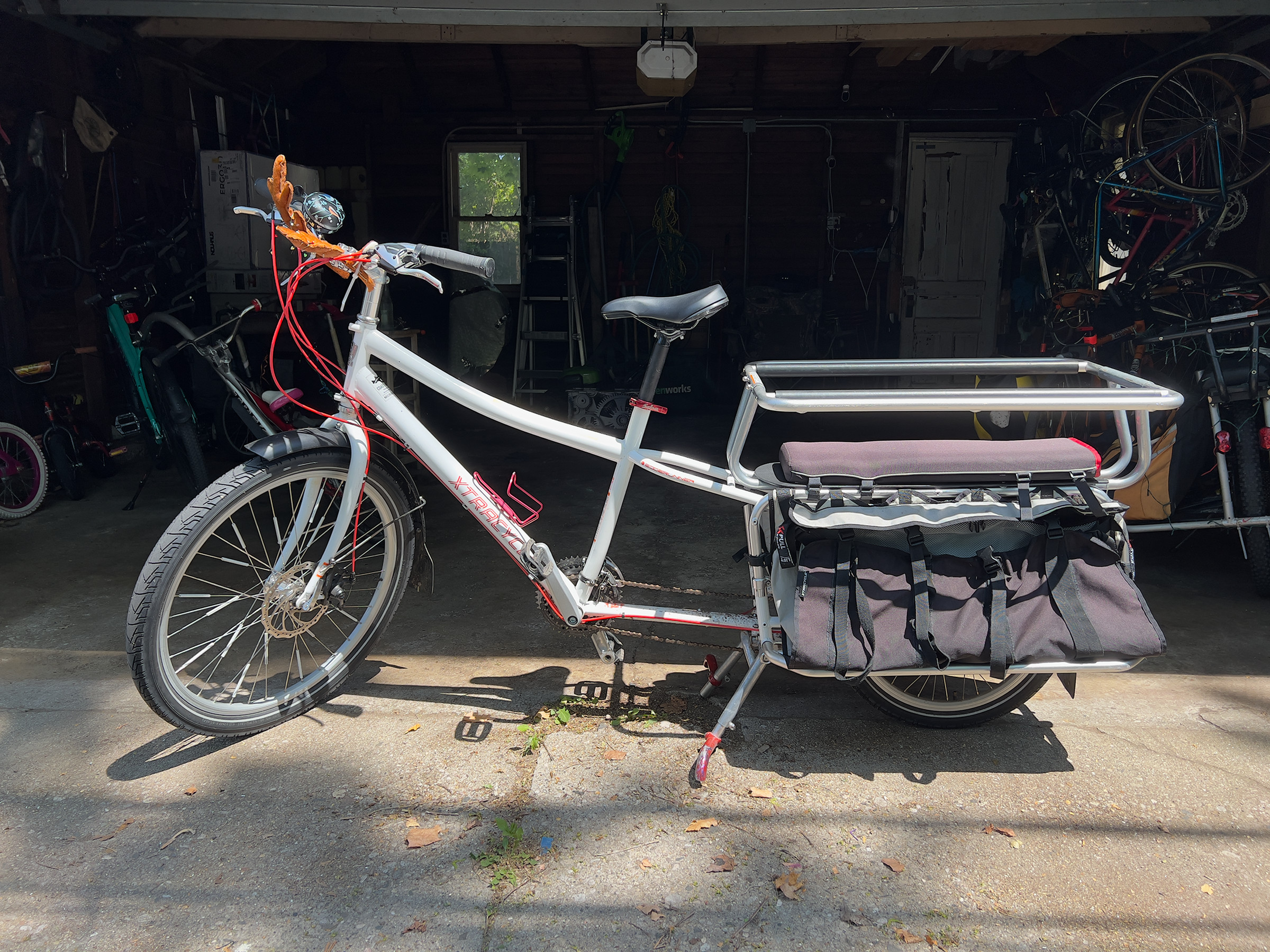 A picture of the Xtracycle Edgerunner without the e-bike motor on it, on its kickstand in front of an open garage. The bike is white with red lettering on the downtube, has a child-carrying modification in the back with a padded seat, and a large bag is visible hanging on the side.