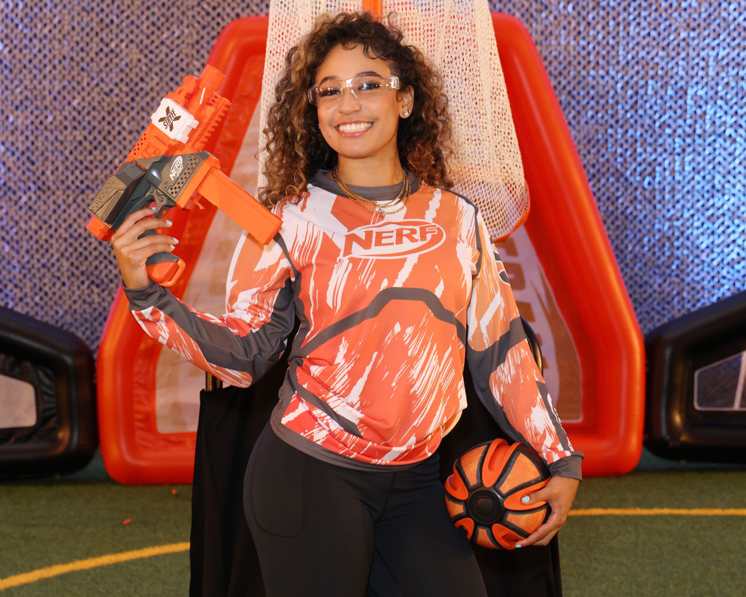 A college athlete poses with the Nerfball and Stryfe X blaster while wearing a hit-detection suit in orange and white.