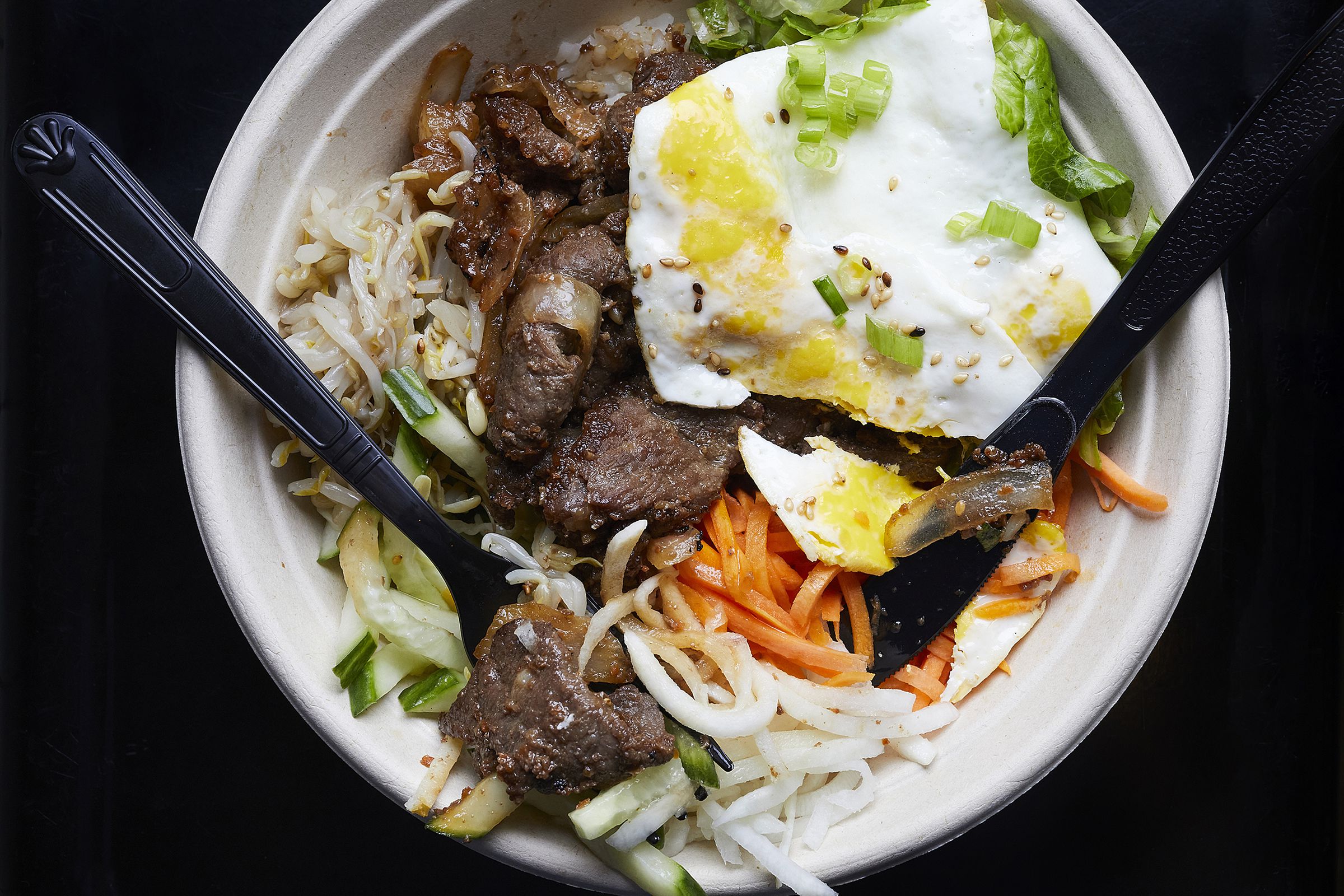 A fried egg and slices of beef lie on a bed of rice and vegetables in a bowl along with a plastic knife and fork.