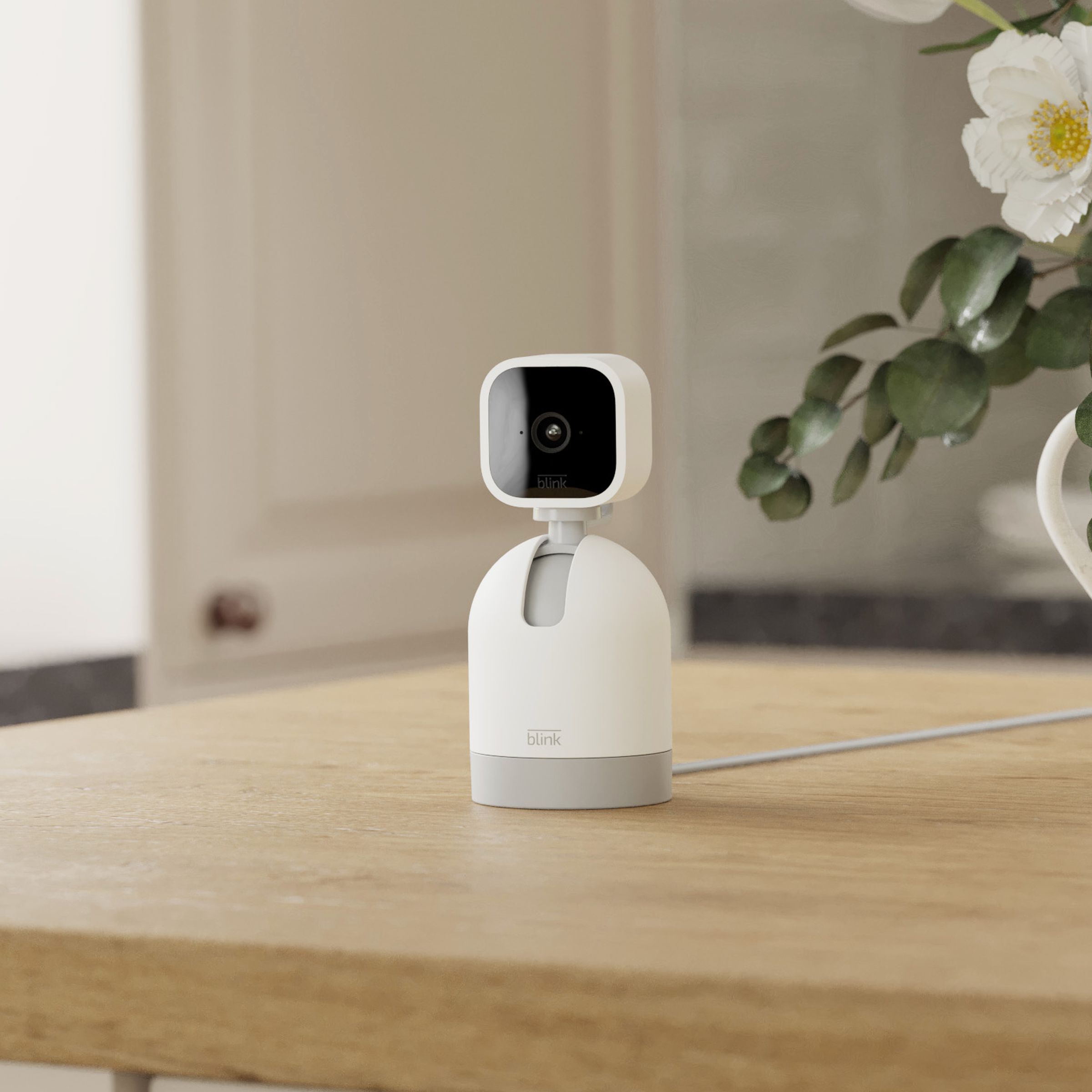 The scene is a wooden kitchen counter with a flower pot offset to the right, and in the center is the Blink Mini cam, which is a blocky black and white device. It’s mounted on a white bullet-shaped body that makes it look like a Pixar-style robot device. There’s a cable running out the back.