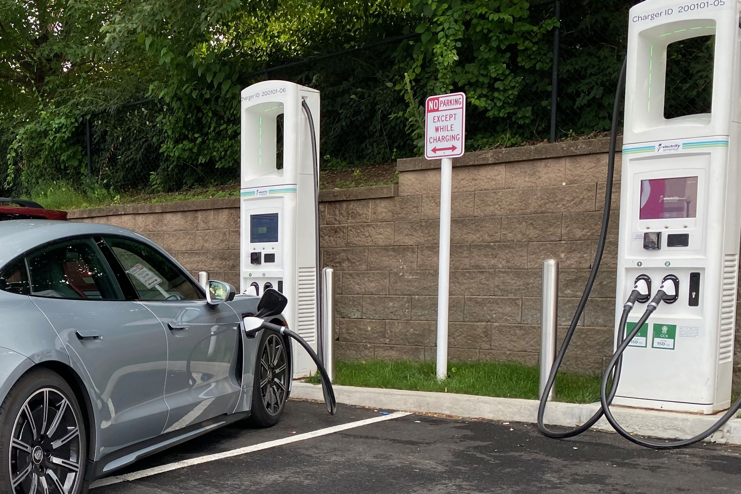 two electrify America stations with two plugs each are divided in stalls with a “no parking except while charging” sign in the middle. A silver Porsche taycan is plugged into one of the chargers and is parked at an angle that takes up parts of two spaces.