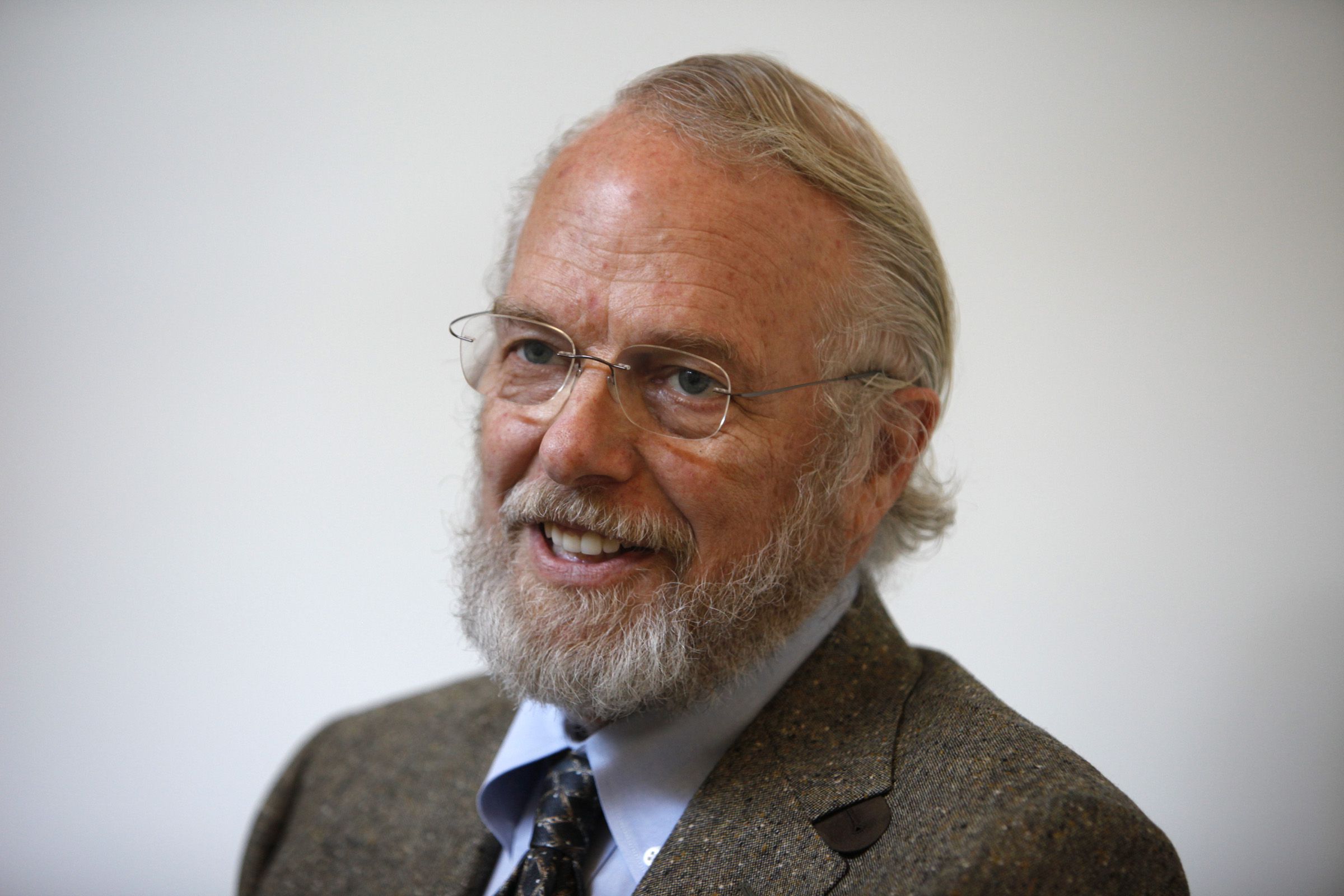 An image of Dr. John Warnock, smiling and looking at someone out of frame. He is wearing glasses, has a short beard, and is wearing a black suit with a blue shirt and black tie.