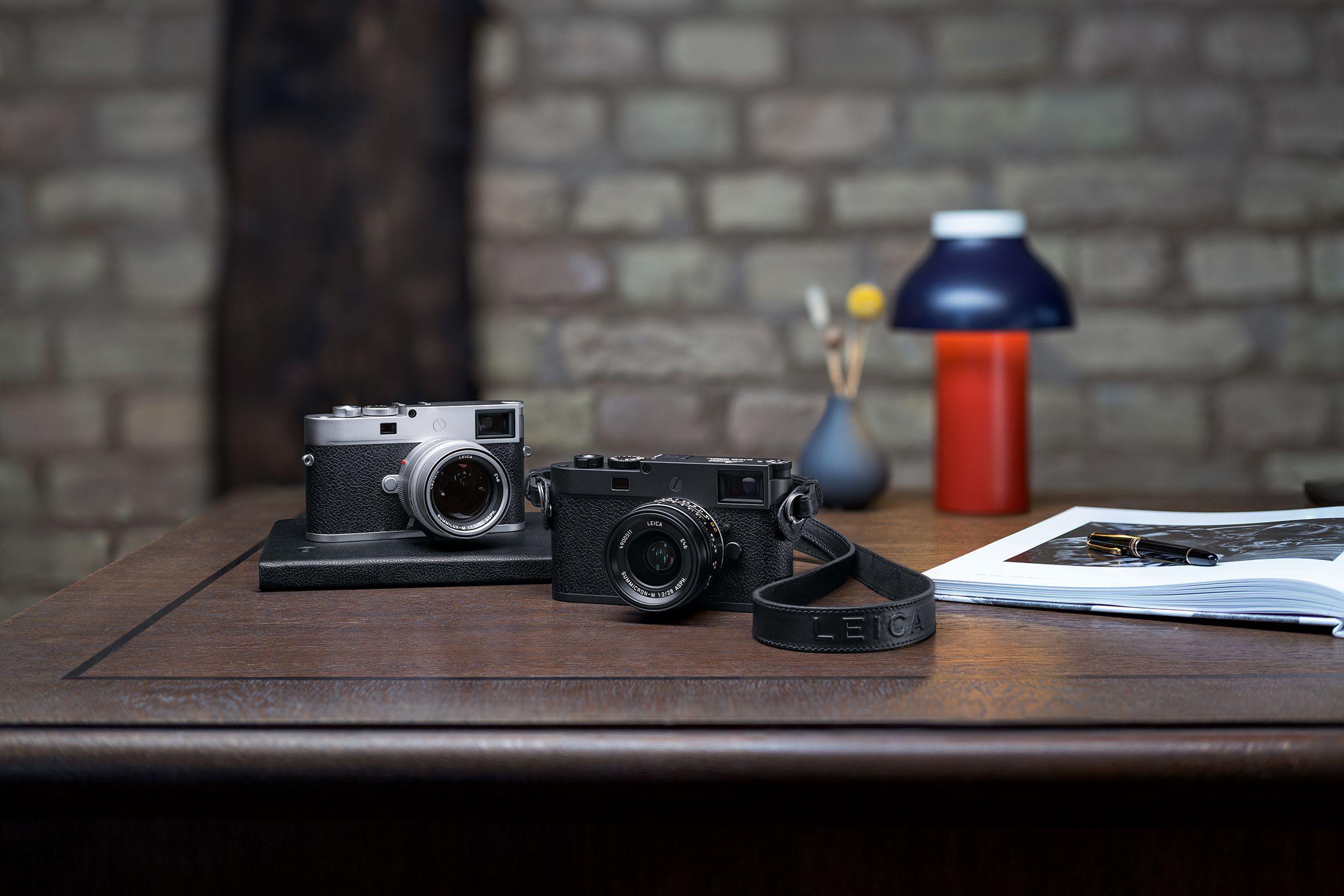 A pair of silver and black Leica M11-P cameras sitting on a wood desk.