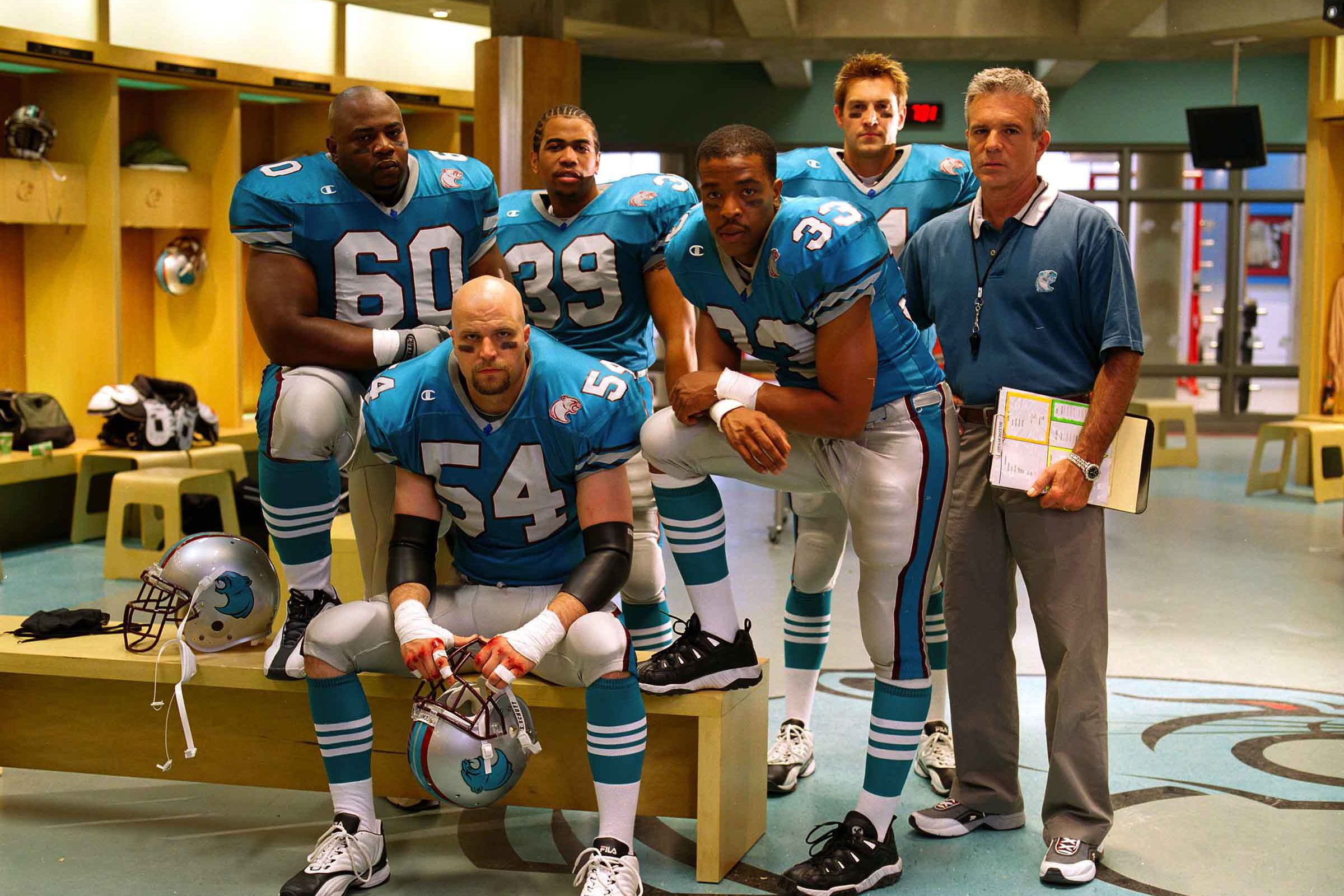 A photo of five Cougars football players in uniform in a locker room. The head coach stands next to them wearing a Cougars polo shirt and holding a clipboard.