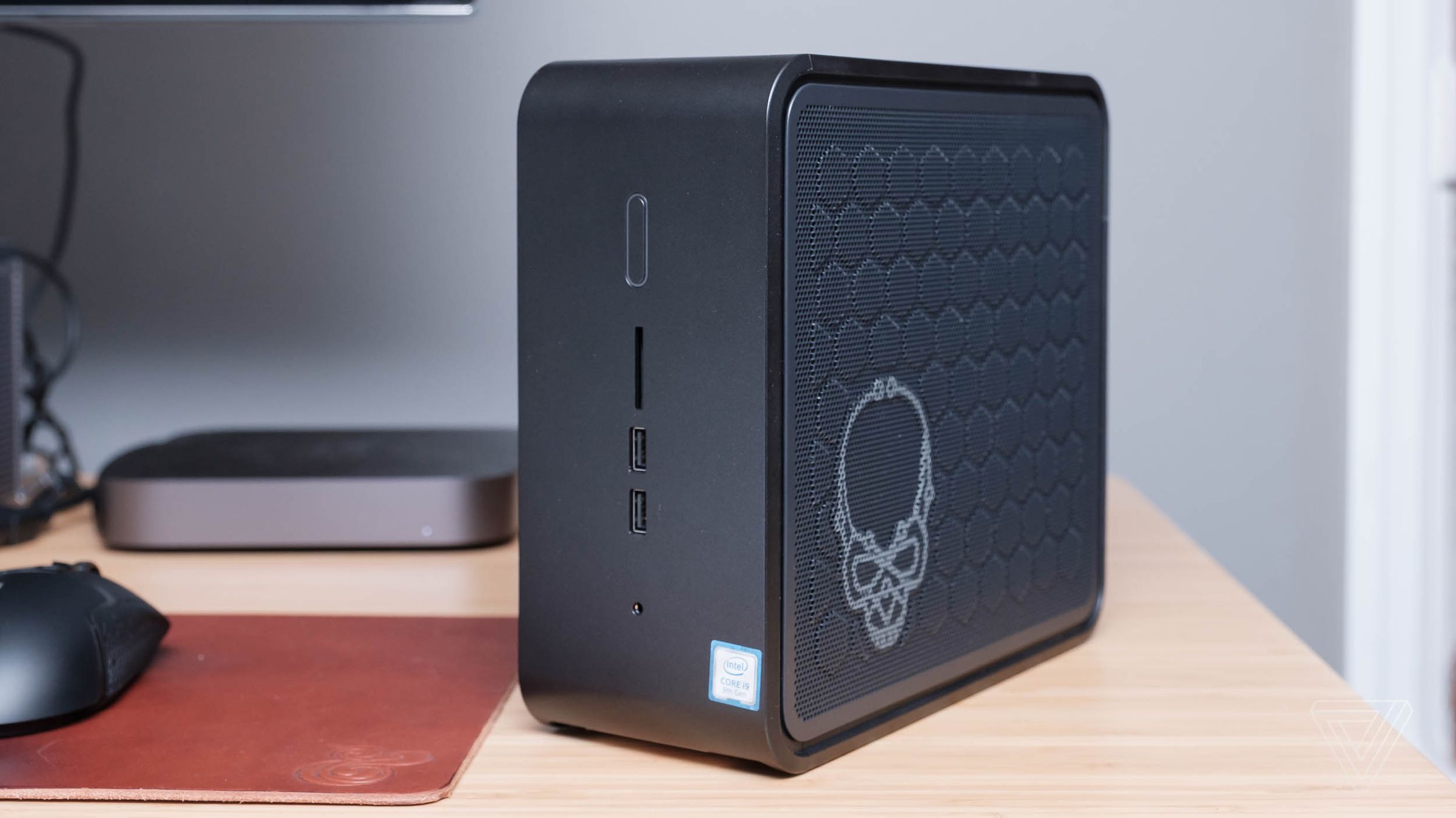 Intel NUC 9 computer standing vertically on a wood desk with a skull etched on the side and a gray Mac Mini in the background